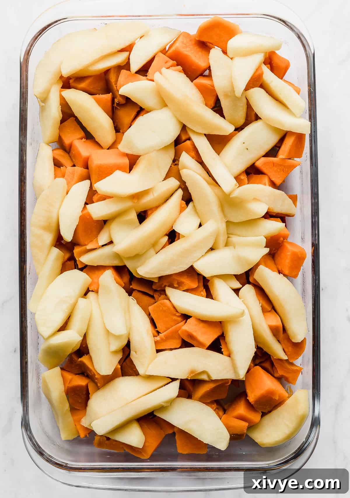 Neatly arranged sliced apples layered over cooked, cubed sweet potatoes in a glass casserole dish, creating a visually appealing foundation.