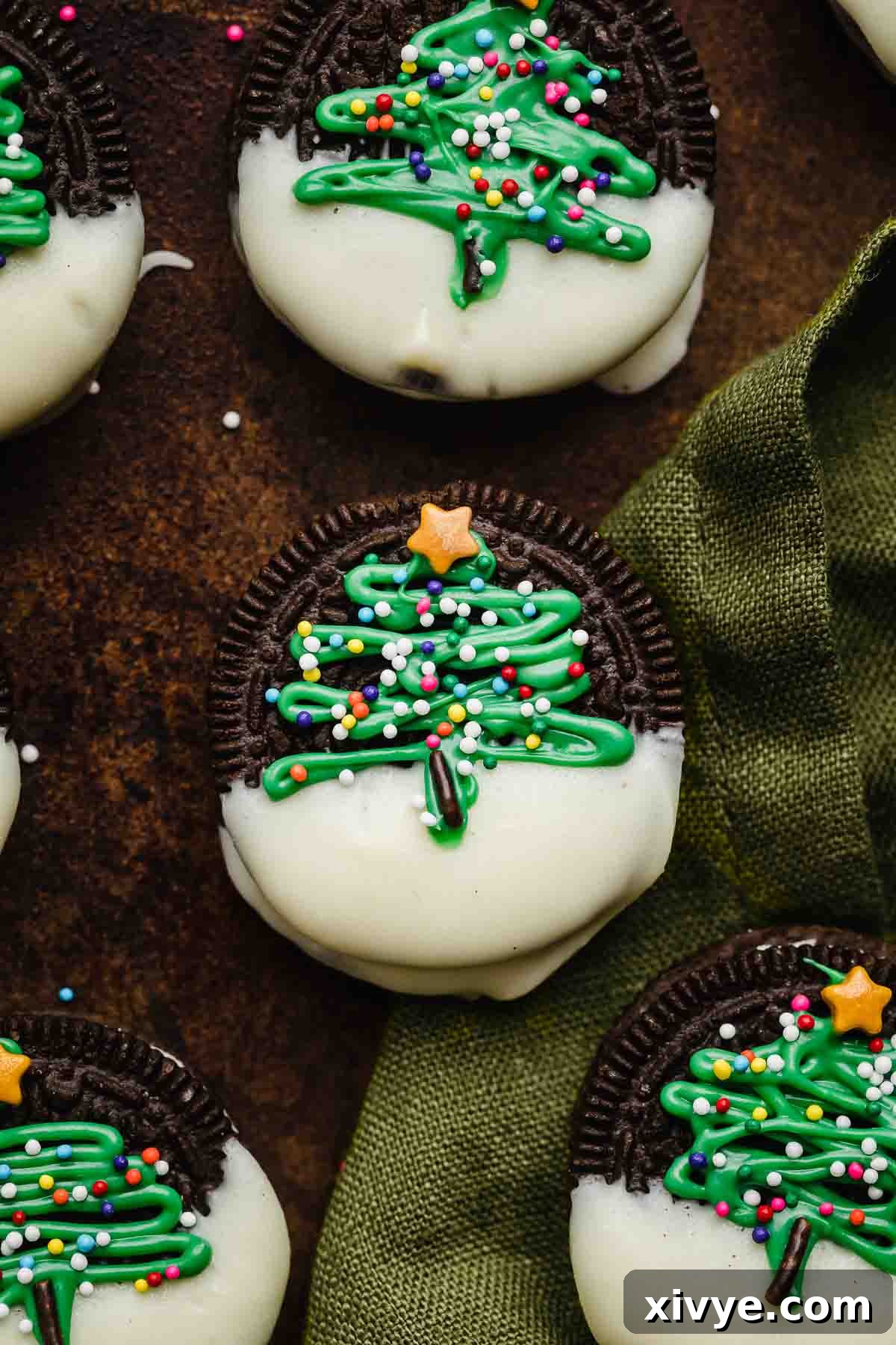 Christmas Oreo on a green linen napkin, the Oreo is half dipped in white chocolate then has a green abstract christmas tree piped on the Oreo and sprinkled with nonpareil sprinkles and a star sprinkle on top.