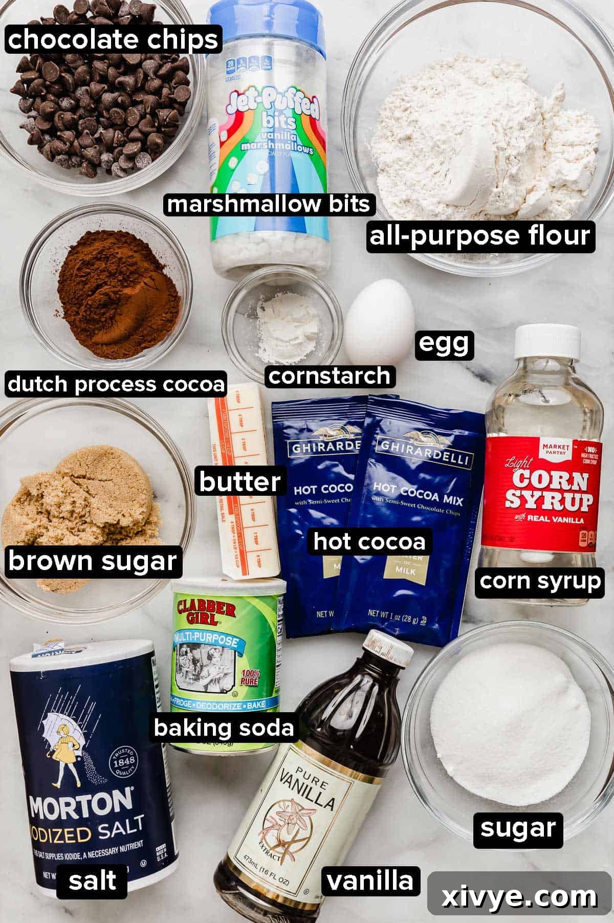 Hot cocoa cookie ingredients on a white marble background: vanilla, salt, flour, chocolate chips, dehydrated marshmallows, hot cocoa mix, sugar, and corn syrup.