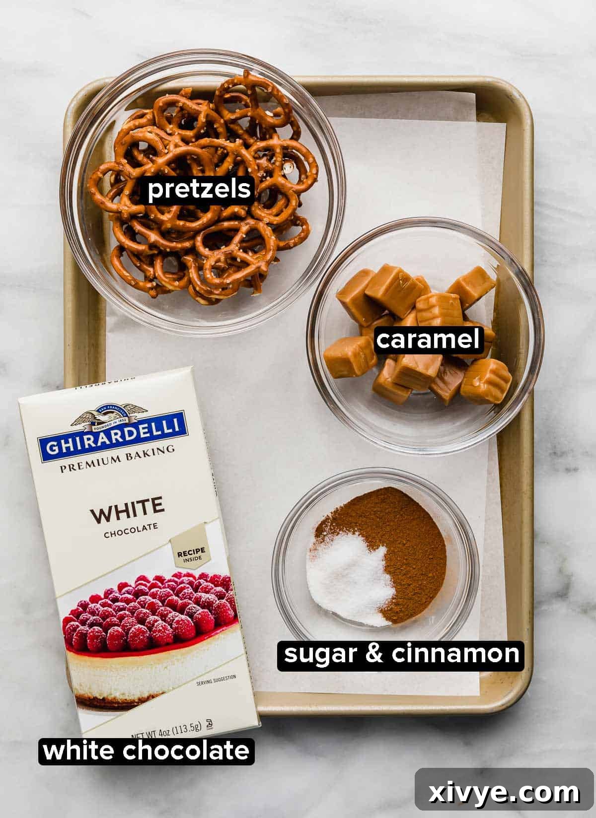 A baking sheet with white chocolate bar, pretzels, caramel squares, and cinnamon sugar, ready for making snickerdoodle pretzels.