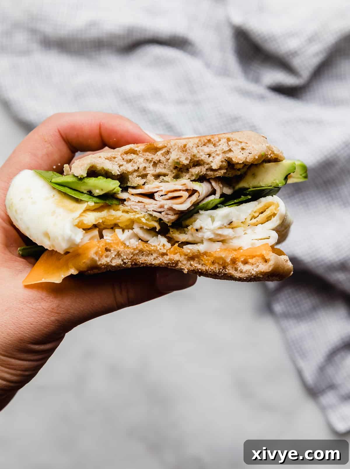 A hand holding a Turkey Breakfast Sandwich on an English muffin topped with avocado, spinach, turkey, eggs, and cheese, ready to be enjoyed.
