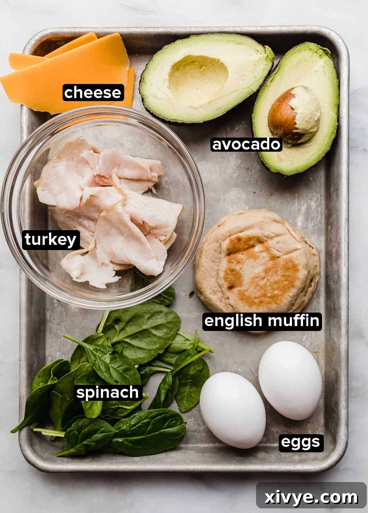 Turkey Breakfast Sandwich ingredients laid out on a baking sheet: slices of deli turkey meat, fresh spinach, a ripe avocado, various cheeses, eggs, and an English muffin.