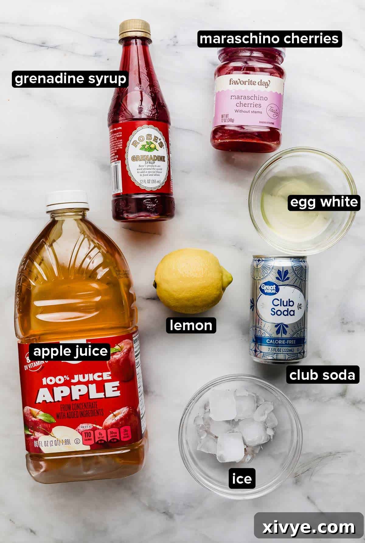 Pink Elegance Sparkler 3 Pink Lady Mocktail ingredients on a white marble background: grenadine syrup, egg white, lemon, apple juice, club soda, ice, and maraschino cherries.