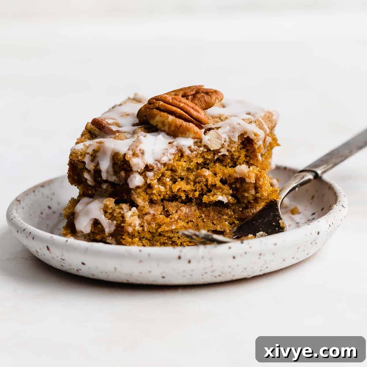 Golden Pumpkin Pecan Crumble Coffee Cake 7 A freshly baked, glazed pumpkin coffee cake slice, adorned with a single pecan half, resting on a white plate against a clean white background.