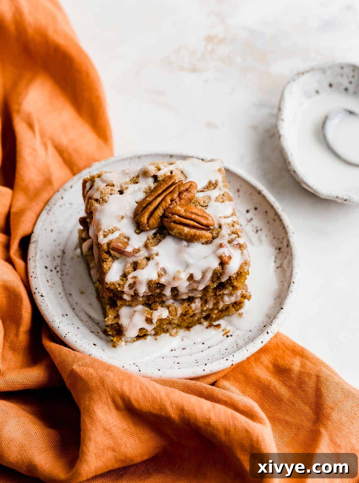 Golden Pumpkin Pecan Crumble Coffee Cake 8 A perfectly portioned slice of glazed pumpkin coffee cake, complete with streusel topping, presented on a white plate atop a textured burnt orange linen napkin.