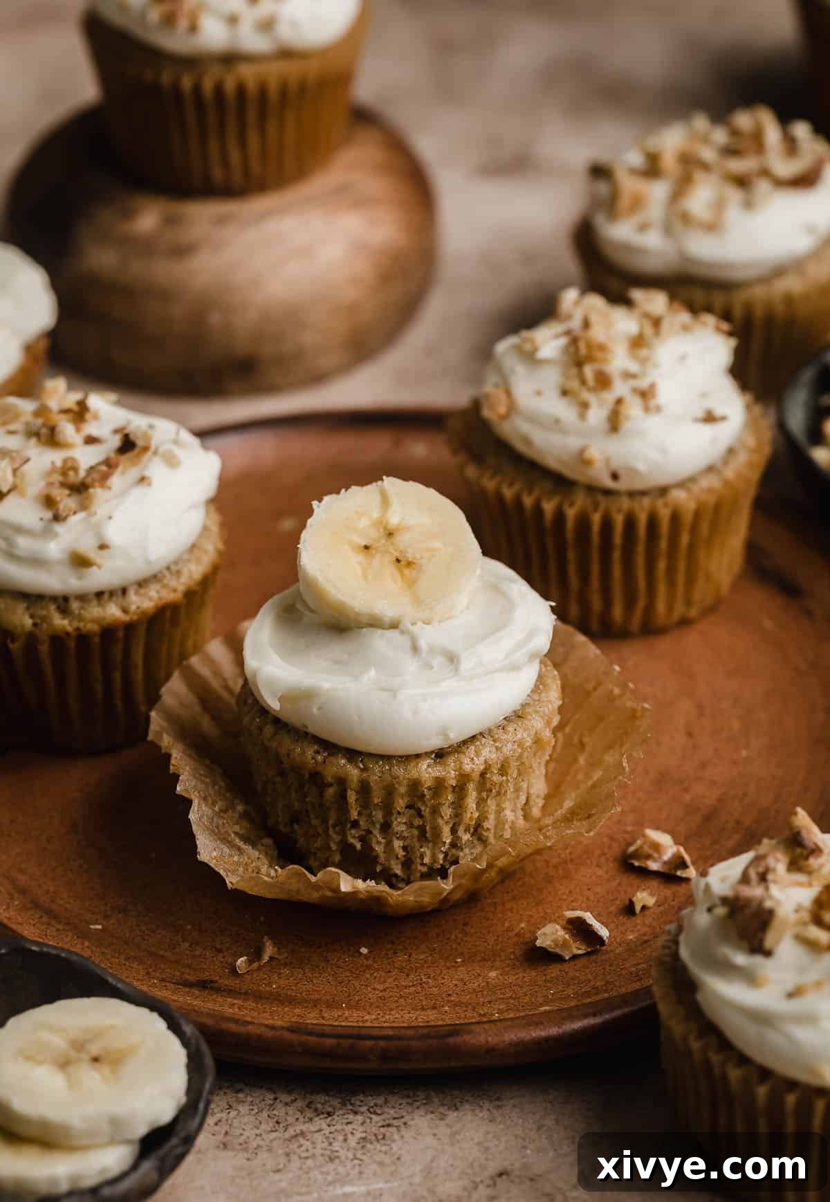 Delicious Homemade Banana Cupcakes 2 Delightful banana cupcakes adorned with creamy cream cheese frosting and a fresh banana slice, elegantly presented on a rustic brown plate.