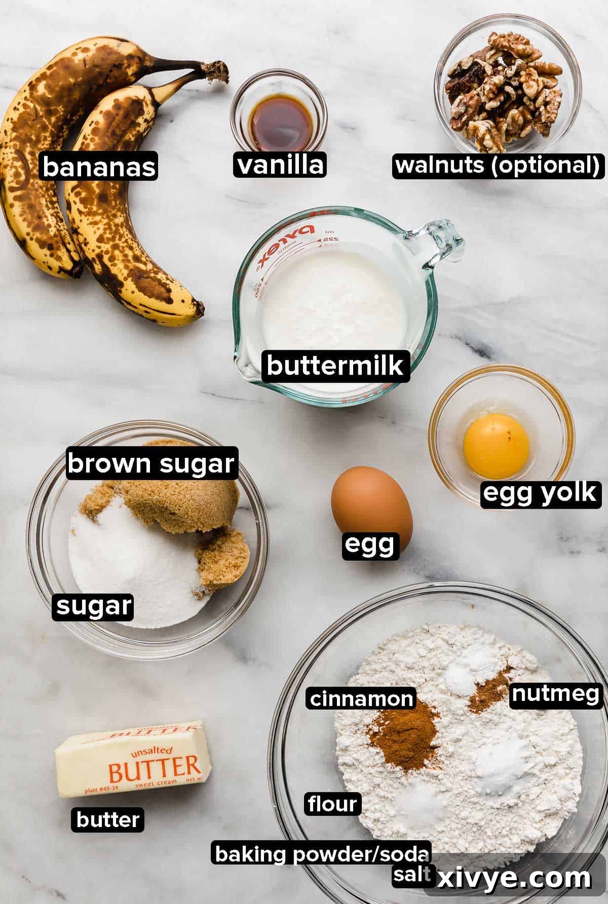 Delicious Homemade Banana Cupcakes 3 A collection of fresh banana cupcake ingredients displayed on a pristine white marble background, featuring perfectly ripe bananas, dry ingredients, eggs, granulated sugar, softened butter, creamy buttermilk, pure vanilla extract, and a scattering of walnuts.