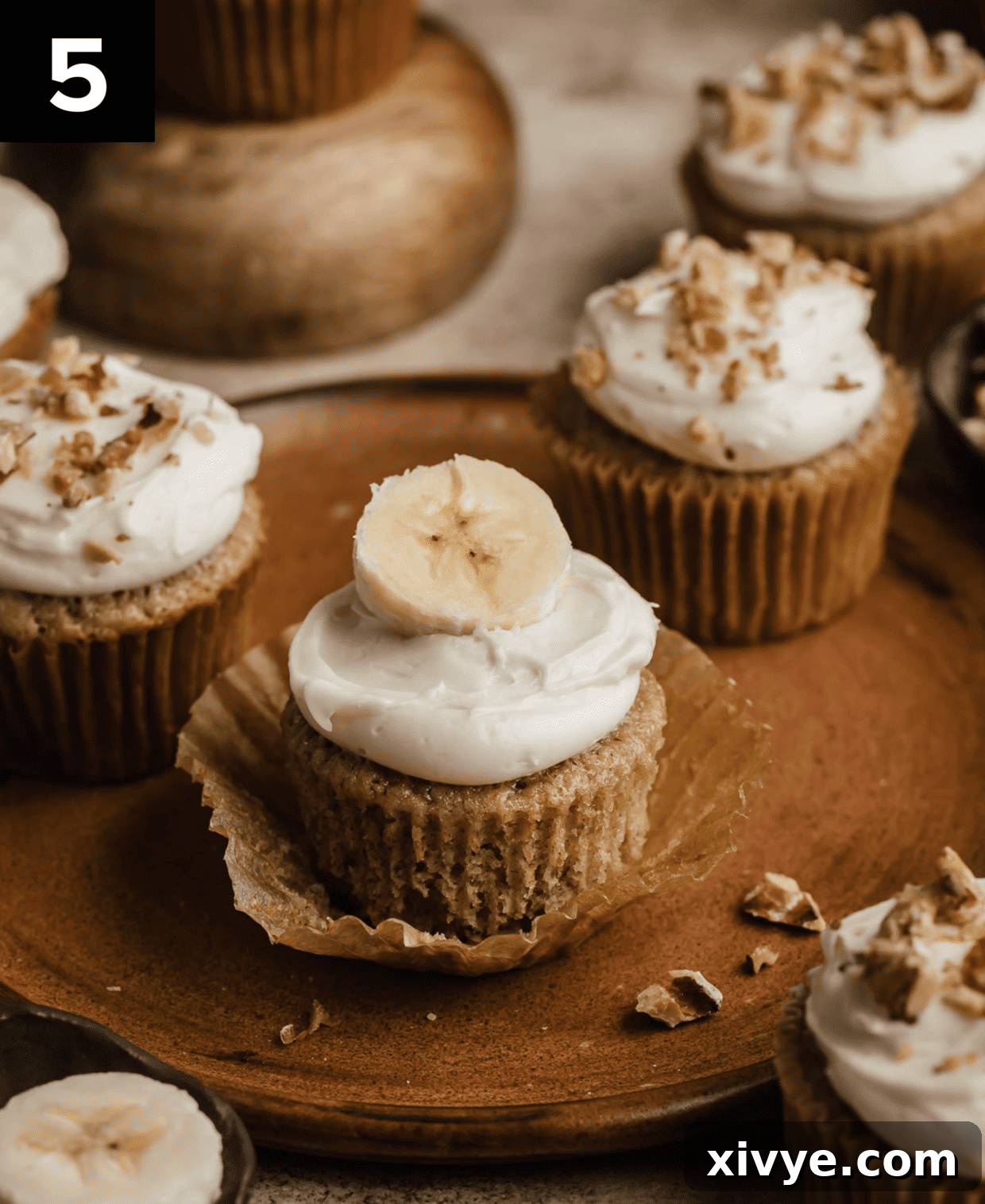 Delicious Homemade Banana Cupcakes 5 A beautifully frosted banana cupcake, crowned with white cream cheese frosting and a delicate fresh banana slice, presented on a charming brown plate.