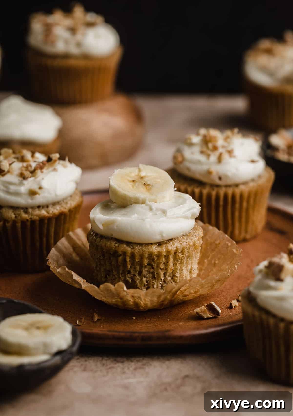 Delicious Homemade Banana Cupcakes 7 A delightful arrangement of banana cupcakes on a brown plate set against a deep brown background, with some cupcakes elegantly topped with a sprinkle of chopped walnuts.