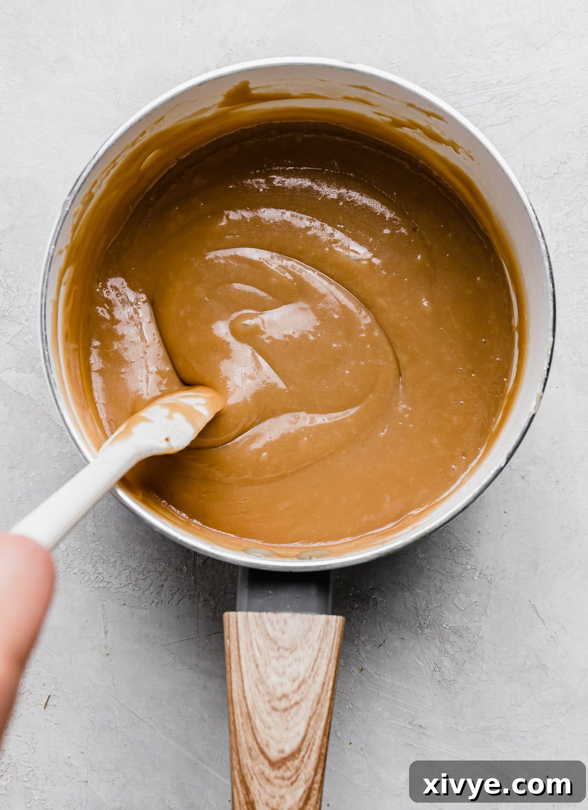 Decadent Banoffee Pie with Crunchy Graham Crust 7 A wooden spoon stirring light colored dulce de leche in a white saucepan, used for an easy banoffee pie recipe.