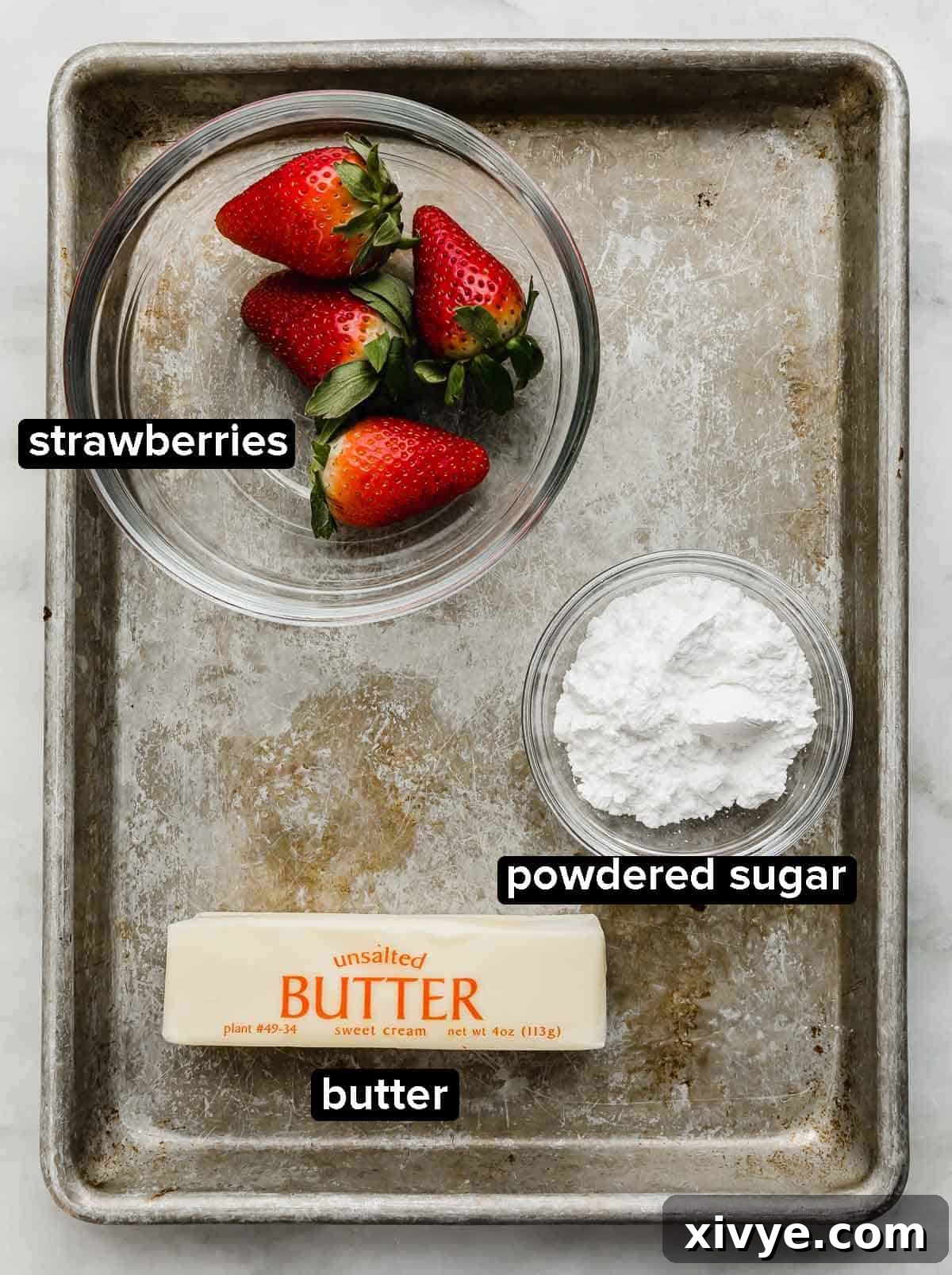 Strawberry Butter ingredients portioned into glass bowls on a metal baking sheet: fresh strawberries, powdered sugar, and a cube of unsalted butter.
