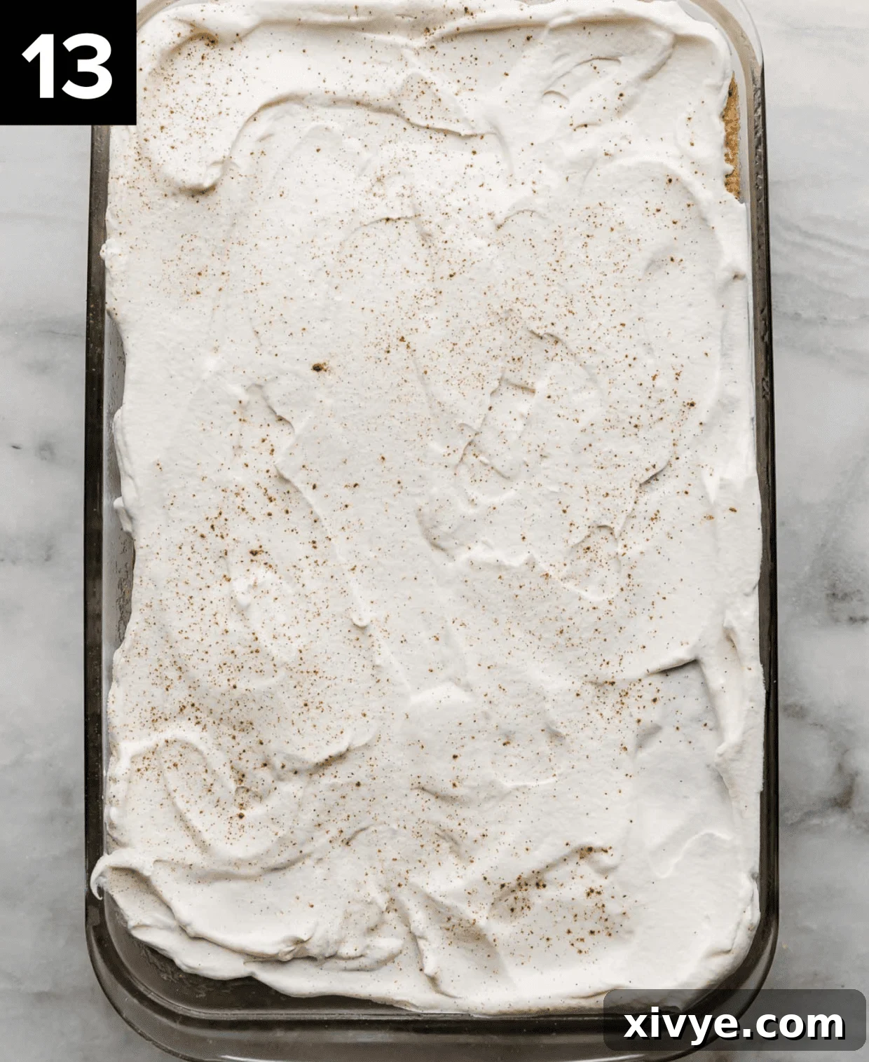 Heavenly Mexican Tres Leches Cake 9 A whipped cream and cinnamon sprinkled tres leches cake in a glass rectangle bowl on a white marble surface.