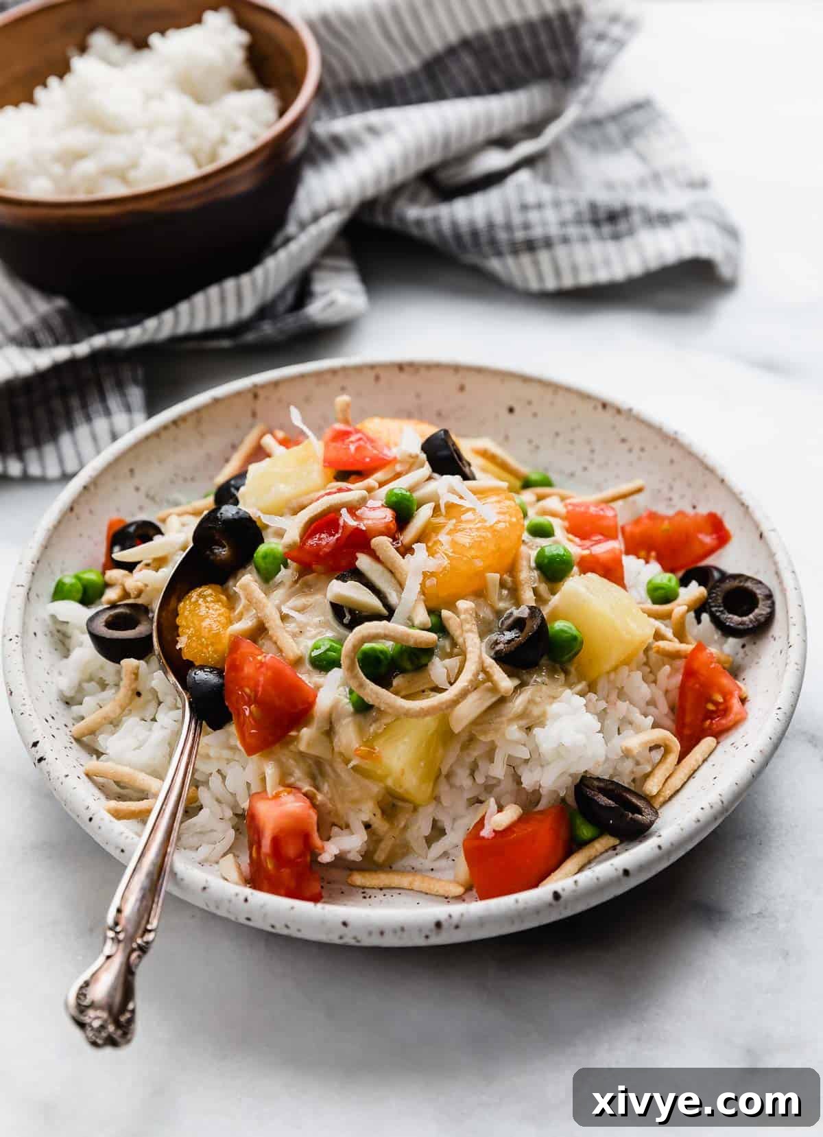 A white plate with a generous serving of white rice and creamy chicken mixture (Crock Pot Hawaiian Haystacks) generously topped with sliced black olives, diced tomatoes, sweet pineapple tidbits, crispy chow mein noodles, and slivered almonds, creating a vibrant and appealing meal.