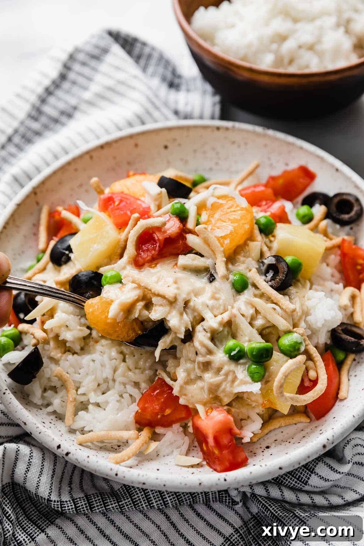 A white plate loaded with the best Hawaiian Haystacks recipe, featuring a mound of creamy chicken and rice, generously topped with vibrant ingredients including diced red tomatoes, sweet pineapple pieces, shredded coconut, black olives, and crunchy chow mein noodles, creating a visually appealing and delicious meal.