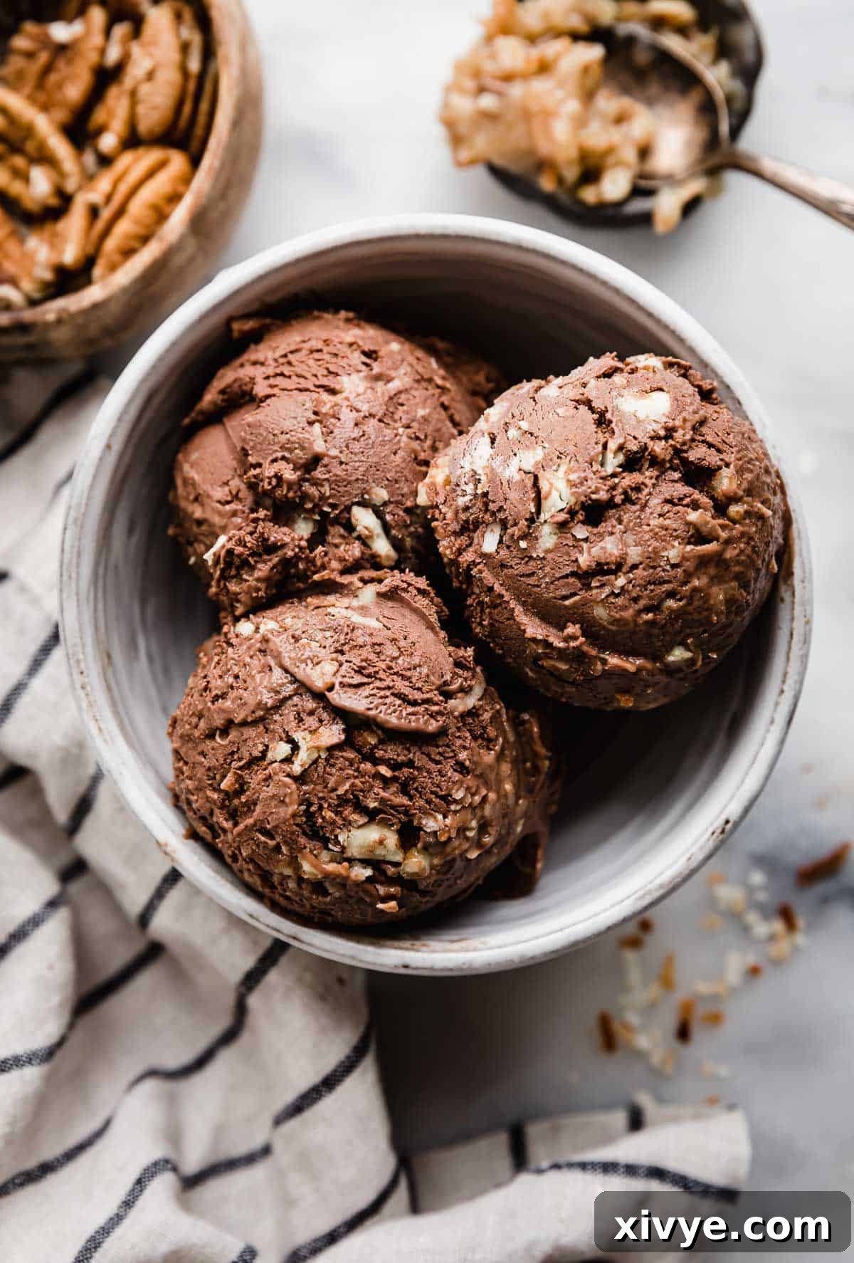 Three scoops of rich dark chocolate German Chocolate ice cream in a pristine white bowl, accompanied by a stylish white and blue stripe napkin.