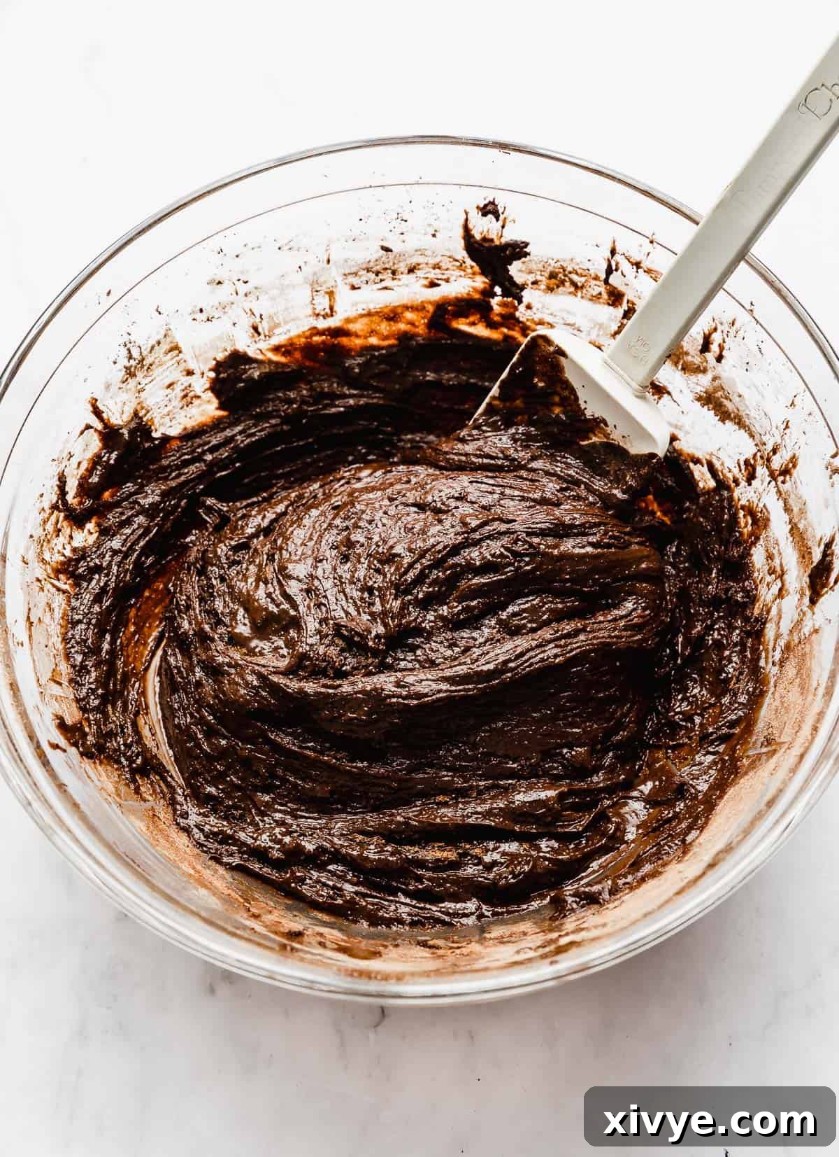 A white spatula stirring the chocolate crinkle cookie batter in a glass bowl, ensuring all ingredients are well-integrated.