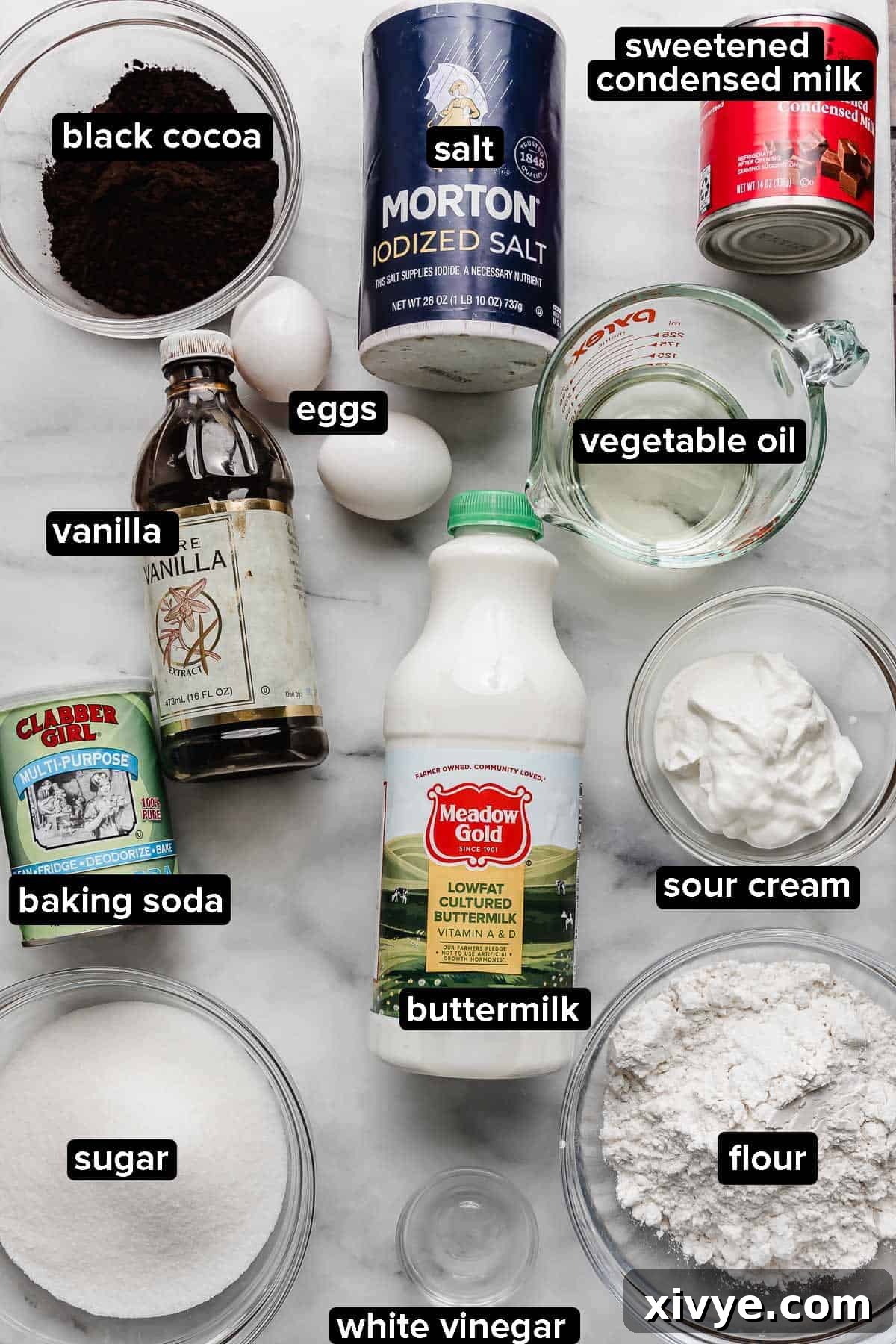 Various ingredients for the Oreo Poke Cake recipe displayed on a white marble surface, including black cocoa powder, buttermilk, sour cream, cake flour, salt, eggs, vegetable oil, and vanilla extract.