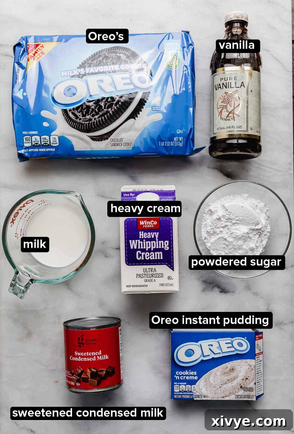Ingredients for the Oreo Poke Cake frosting arranged on a white surface: a package of Oreo cookies, vanilla extract, heavy cream, milk, Oreo instant pudding mix, sweetened condensed milk, and powdered sugar.