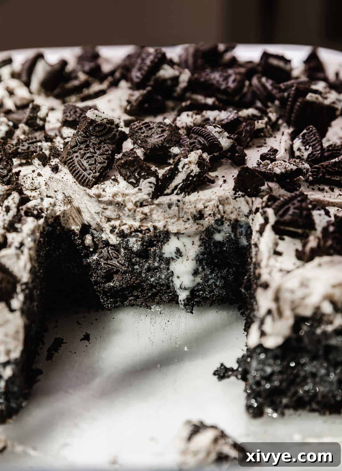 A close-up of an Oreo Poke Cake slice, showing the moist black cake base infused with sweetened condensed milk, topped with fluffy Oreo frosting and crushed Oreos.