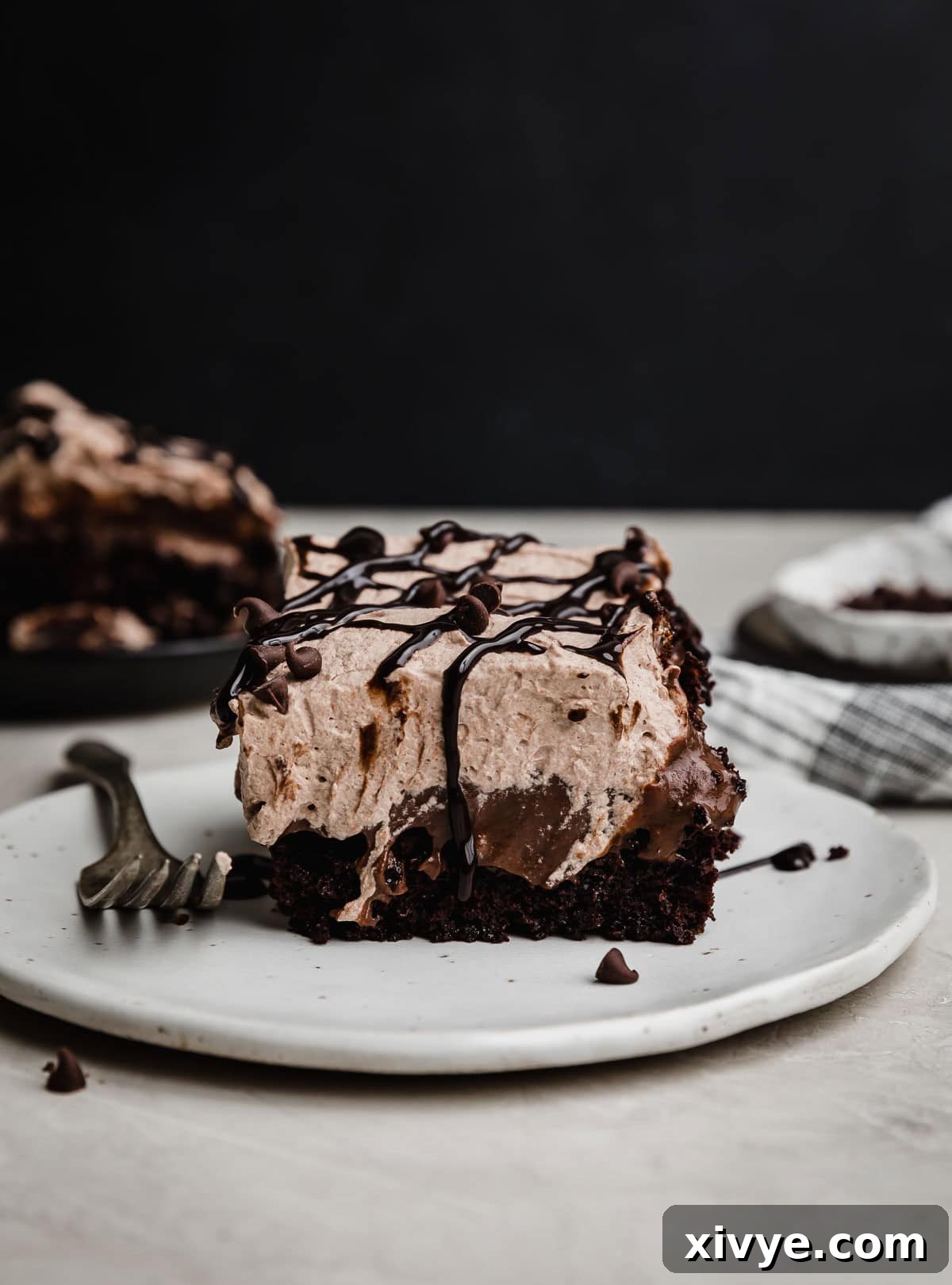 A Chocolate Poke Cake with chocolate pudding and chocolate whipped cream topping on a white plate against a black background.