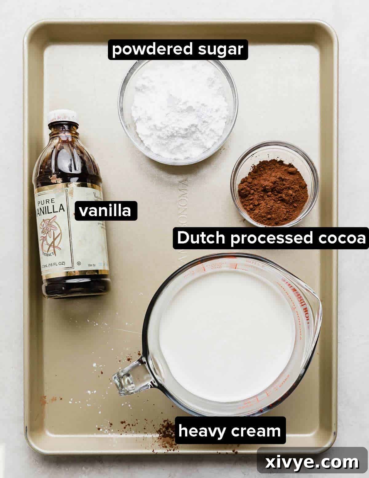 Chocolate Poke Cake ingredients on a gold baking sheet, the ingredients for making the chocolate whipped cream: heavy cream, vanilla, dutch processed cocoa, and powdered sugar.