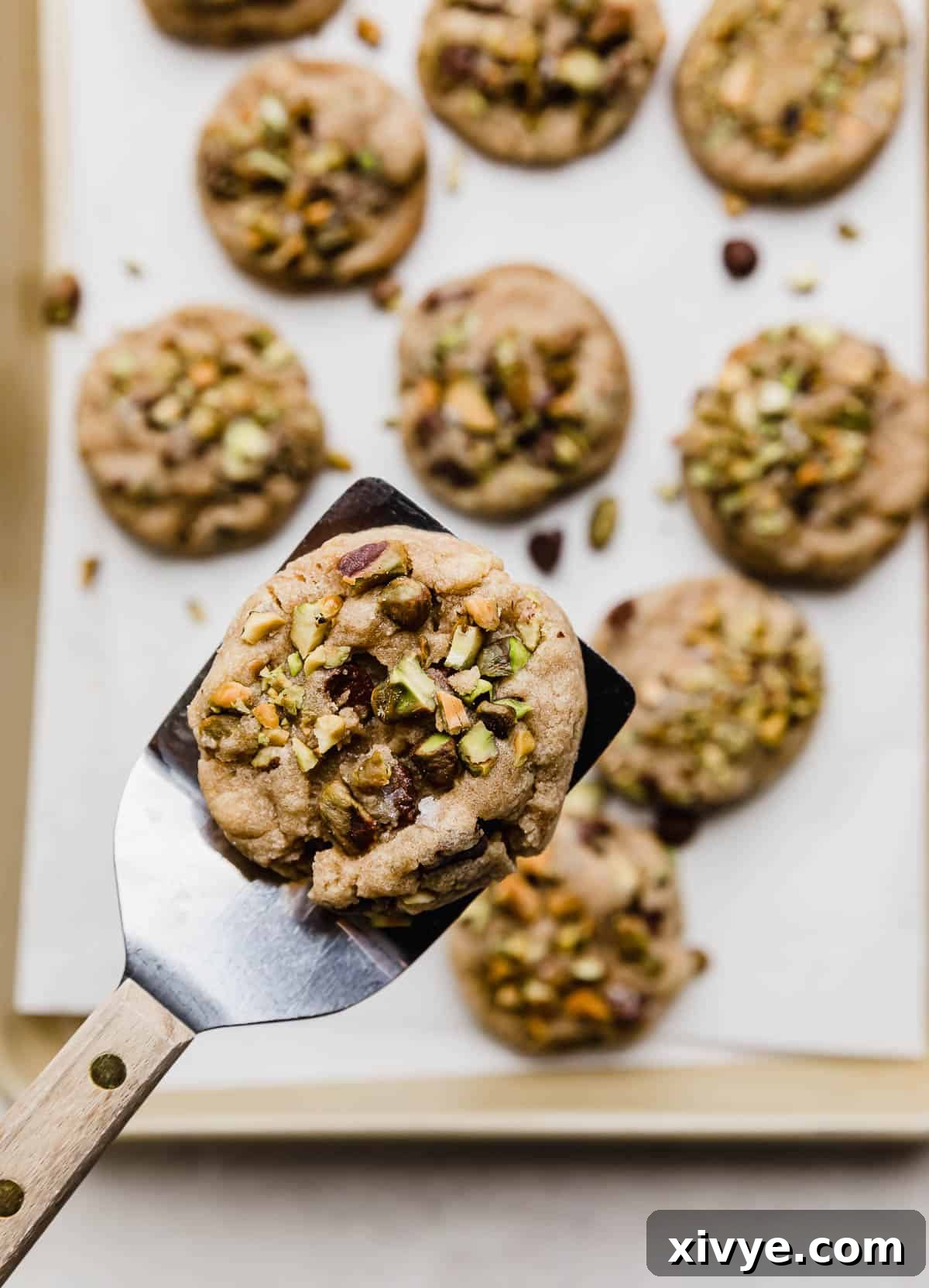 Emerald Pistachio Chocolate Chip Cookies 6 A Chocolate Chip Pistachio Cookie balancing on a metal spatula hovering over a baking sheet of the best pistachio chocolate chip cookies.