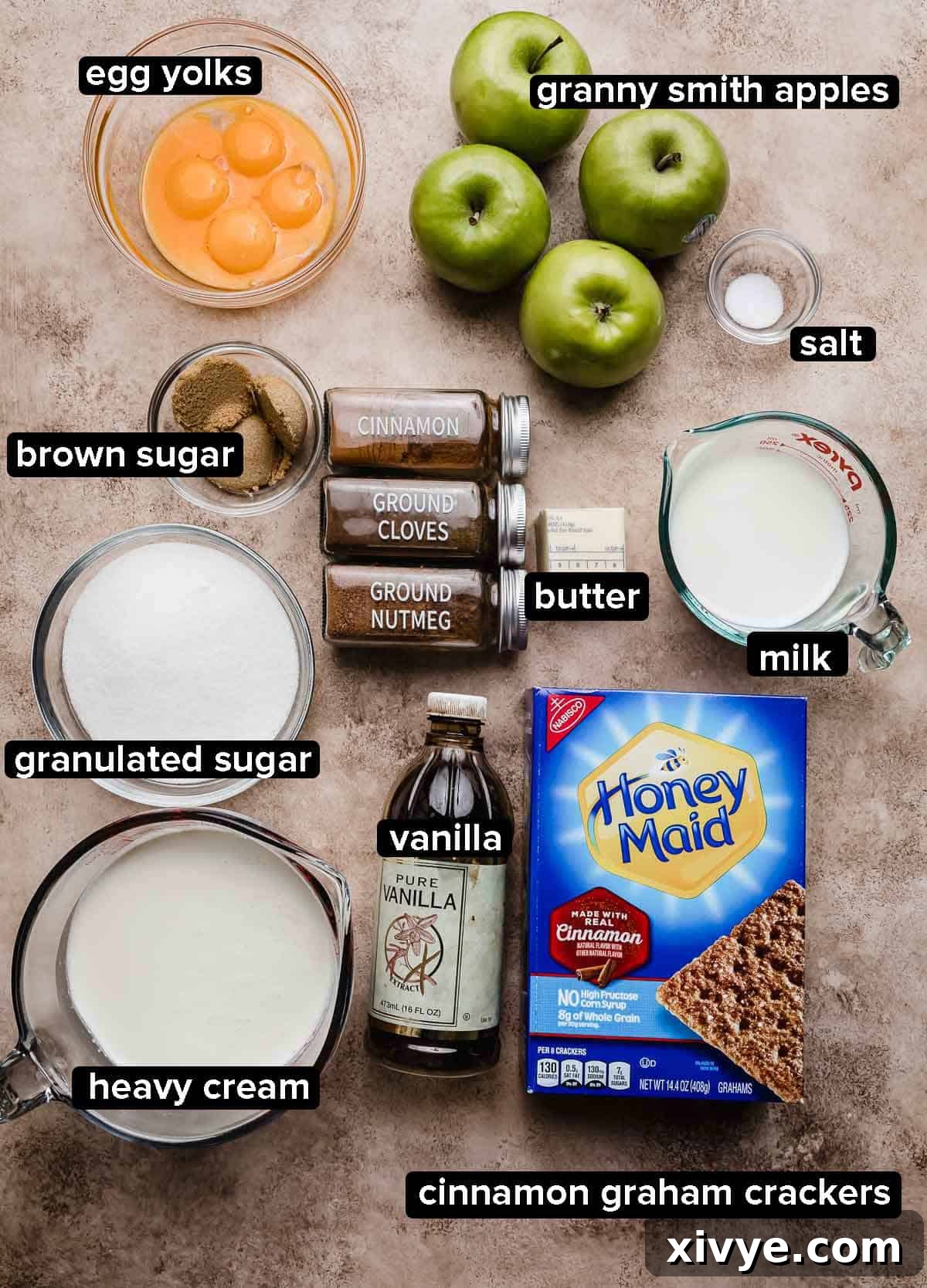 Apple Pie Ice Cream recipe ingredients on a light brown textured surface: Granny Smith apples, cinnamon, heavy cream, egg yolks, sugar, spices, and cinnamon graham crackers.