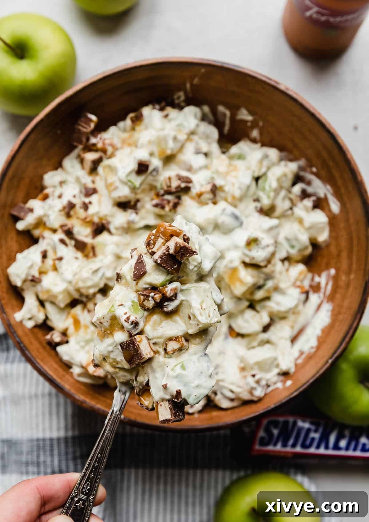 Snickers Apple Delight 2 A brown bowl filled with salad with apples and snickers and topped with caramel, on a white surface.