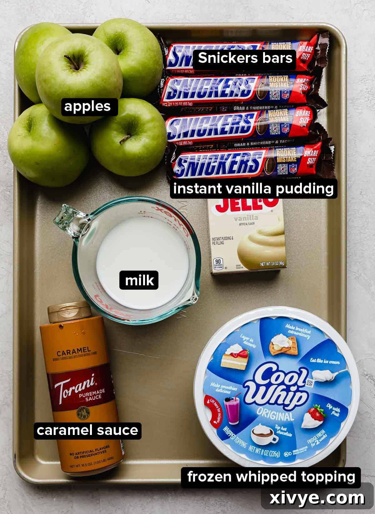 Snickers Apple Delight 3 Apple Snickers Salad ingredients on a baking sheet: snickers, cool whip, milk, vanilla pudding, apples, and caramel.