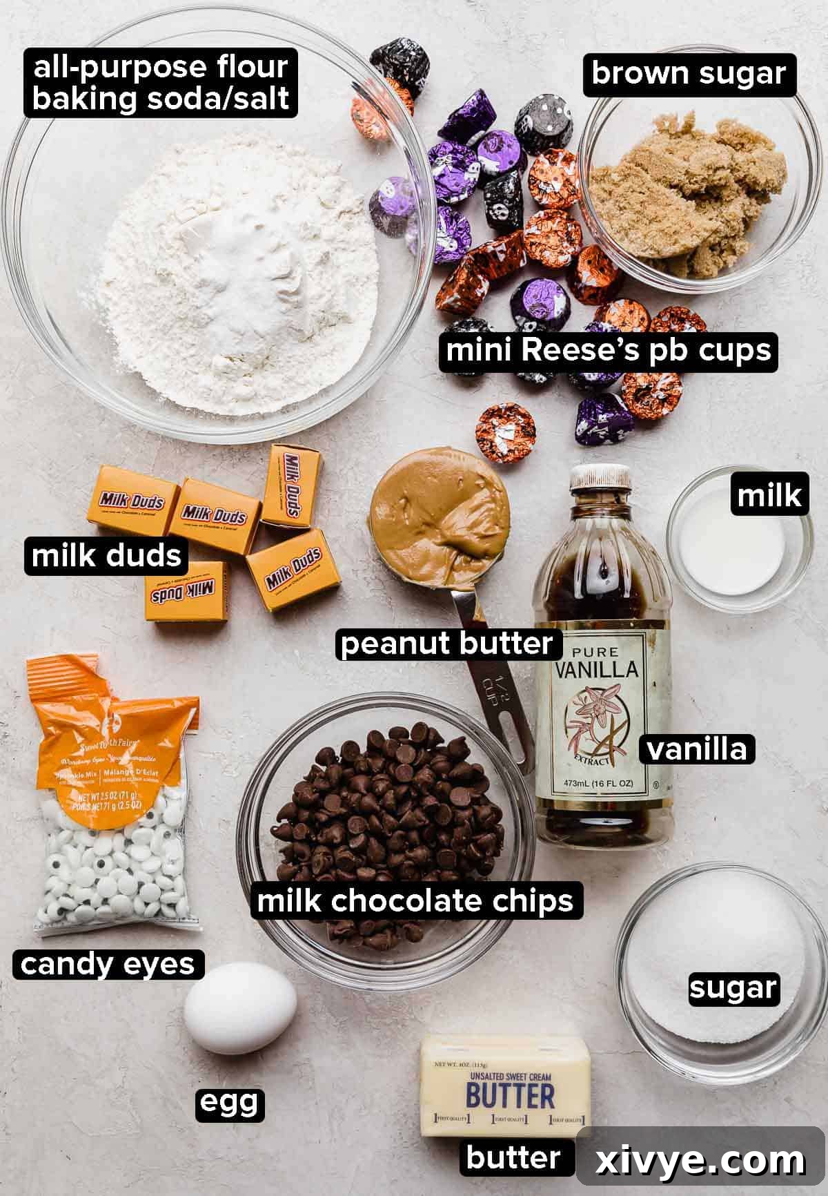 All the ingredients for Halloween Spider Cookies laid out on a light gray surface, including Milk Duds, butter, Reese's peanut butter cups, vanilla extract, creamy peanut butter, sugars, and dry ingredients like flour.