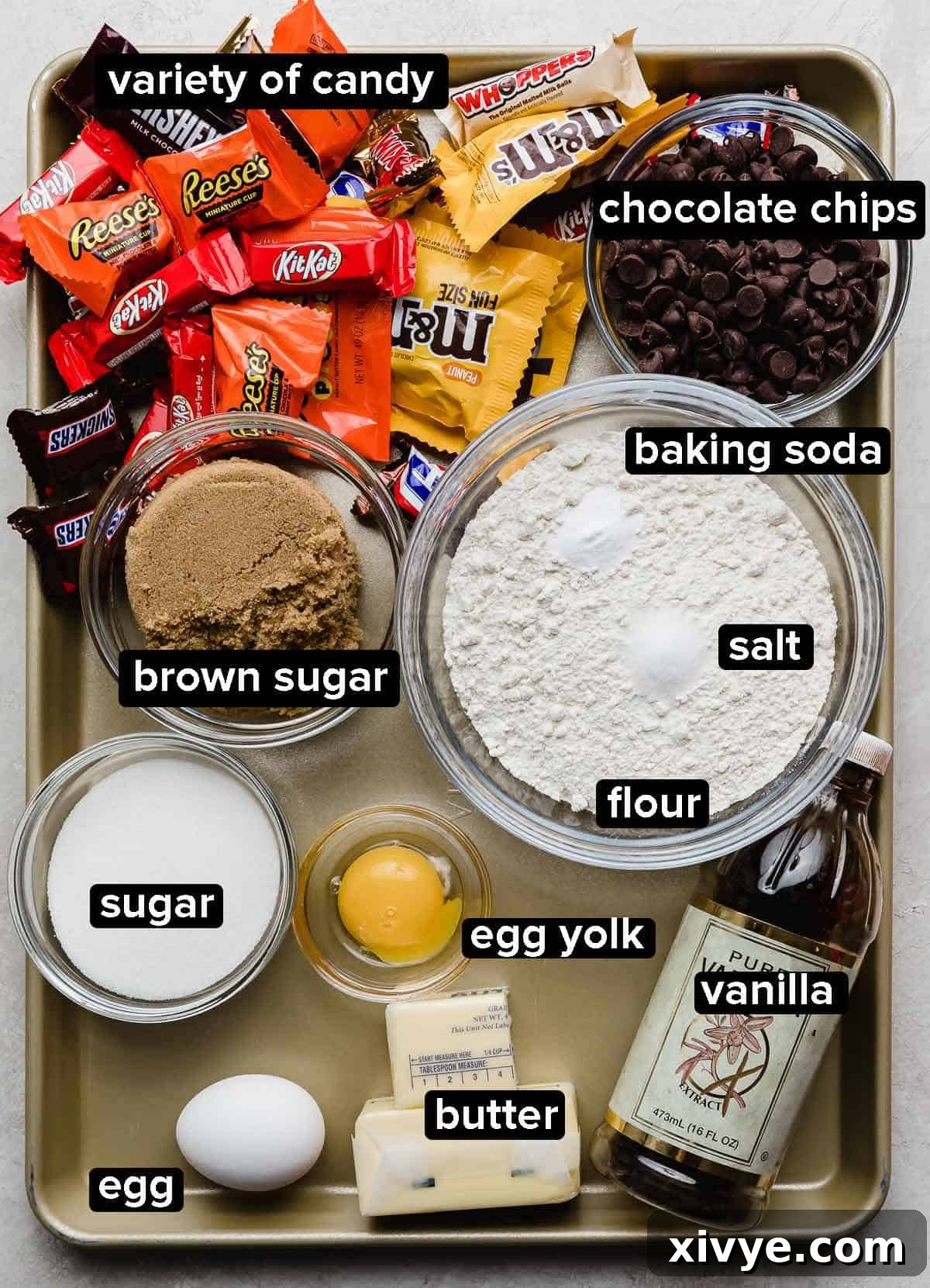 Monster Mash Candy Cookie Bars 3 Leftover Halloween Candy Cookie Bar recipe ingredients portioned into glass bowls on a light gold colored baking sheet: flour, eggs, butter, vanilla, sugar, chocolate chips, and a variety of candy, ready for mixing.