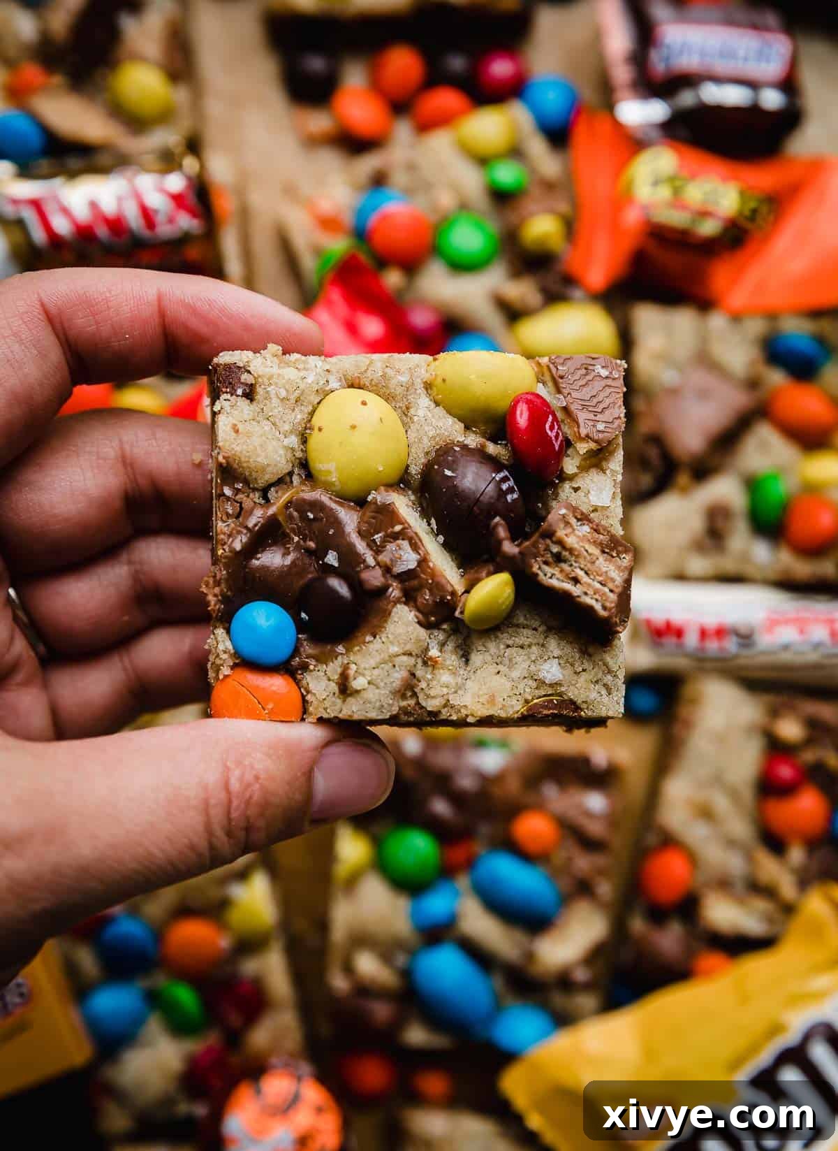 Monster Mash Candy Cookie Bars 6 A hand holding a square piece of Leftover Halloween Candy Cookie Bars topped with M&M's, Milk Duds, and other leftover Halloween candy pieces, showcasing the deliciousness.