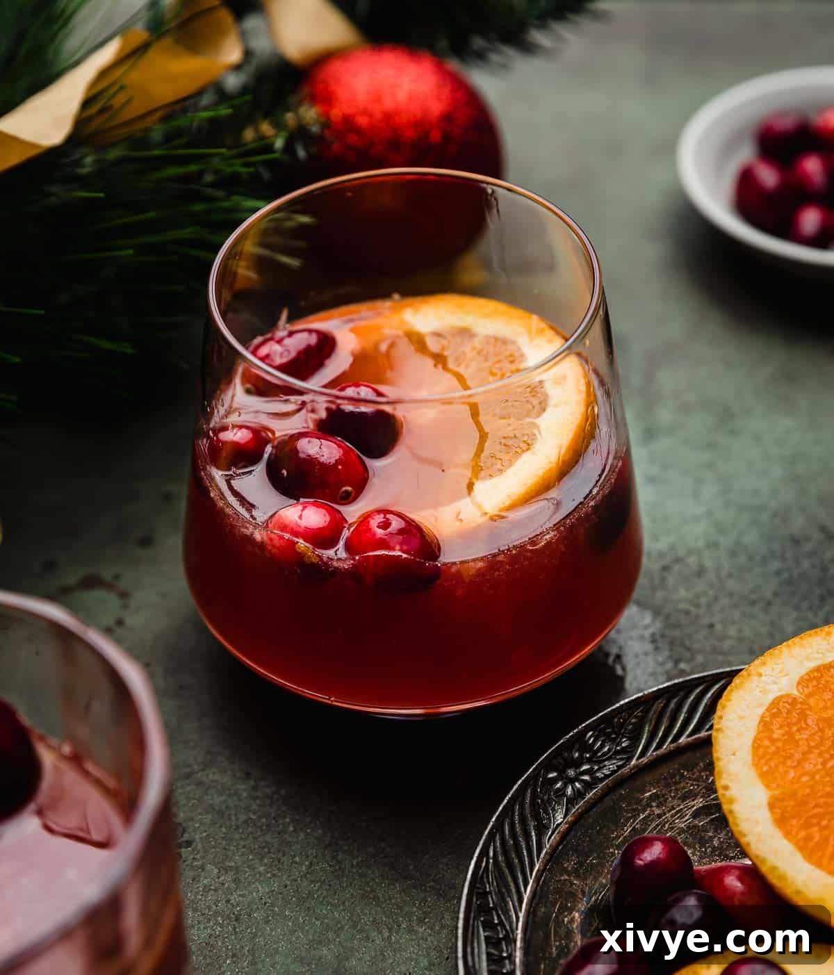 Ruby Refresher 2 A Cranberry orange Mocktail in a glass cup with fresh cranberries and an orange slice, on a dark green surface.