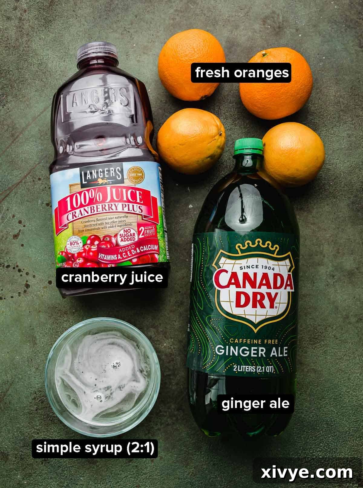Ruby Refresher 3 Cranberry Mocktail ingredients on a green surface: ginger ale, cranberry juice, simple syrup, and fresh oranges.