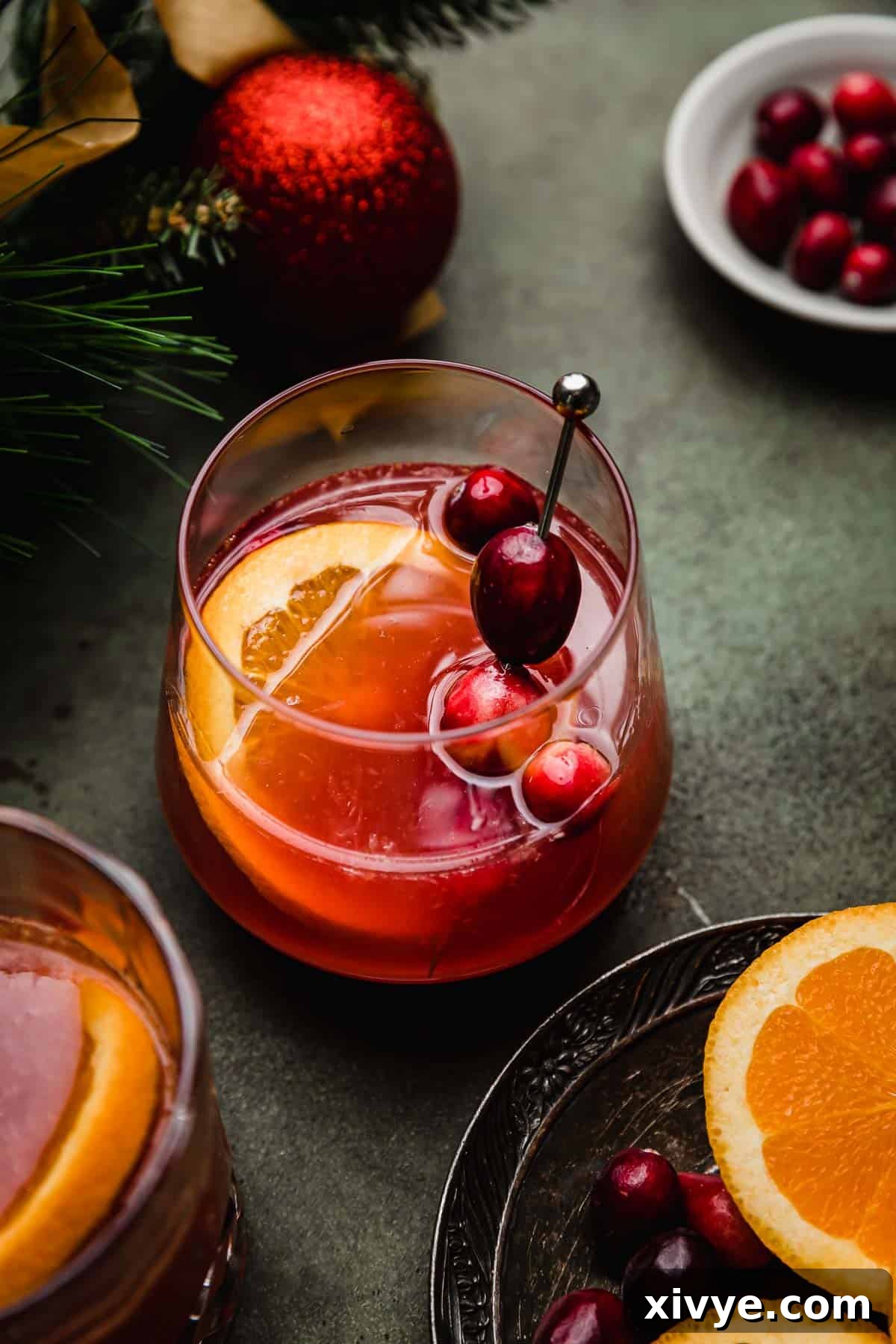 Ruby Refresher 5 A cranberry orange mocktail in a glass cup on a green surface with a slice of orange and fresh cranberries garnishing the drink.