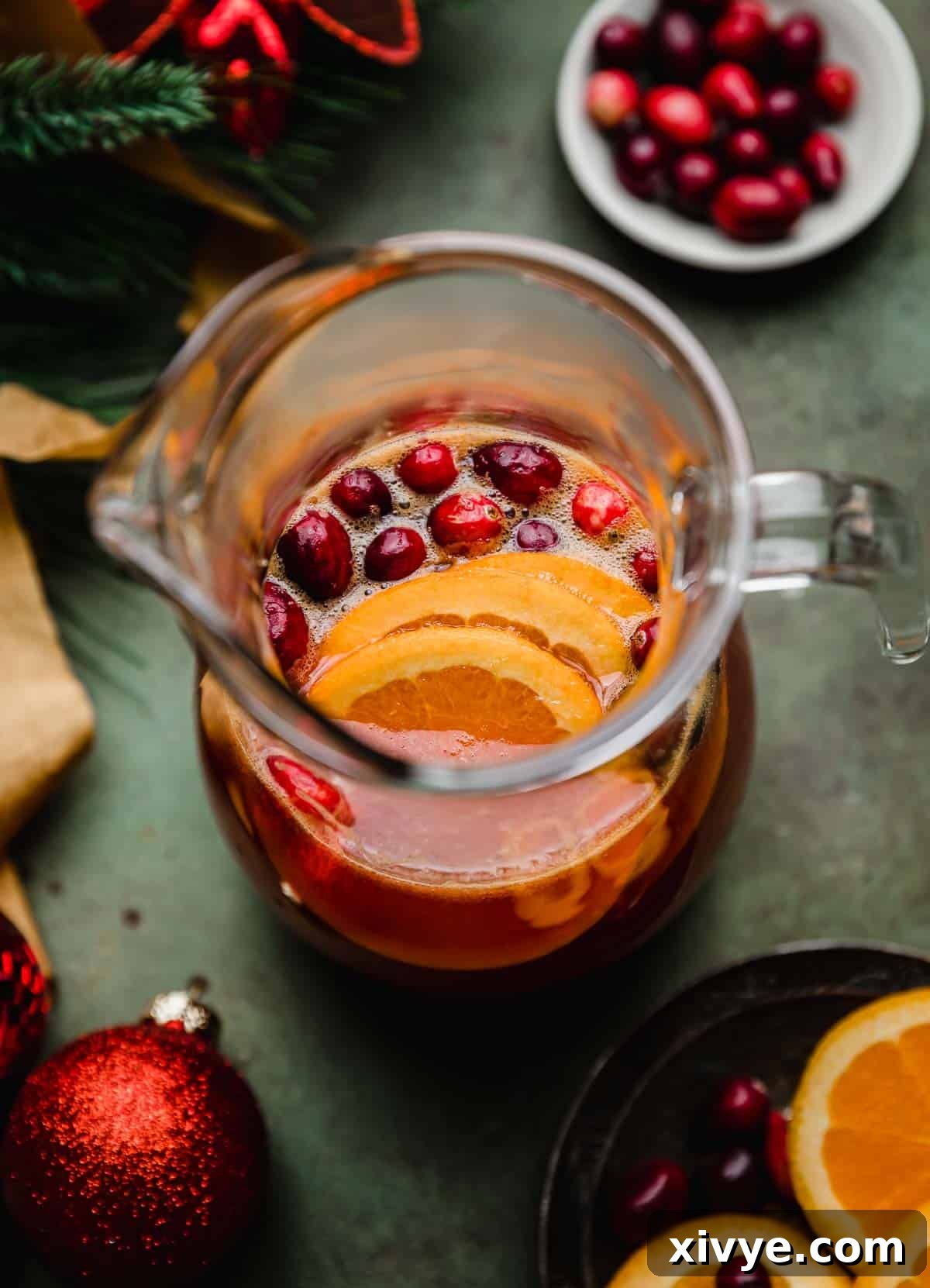Ruby Refresher 6 A cranberry orange mocktail recipe in a glass pitcher on a forest green background.