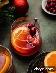 A cranberry orange mocktail in a glass cup on a green surface with a slice of orange and fresh cranberries garnishing the drink.