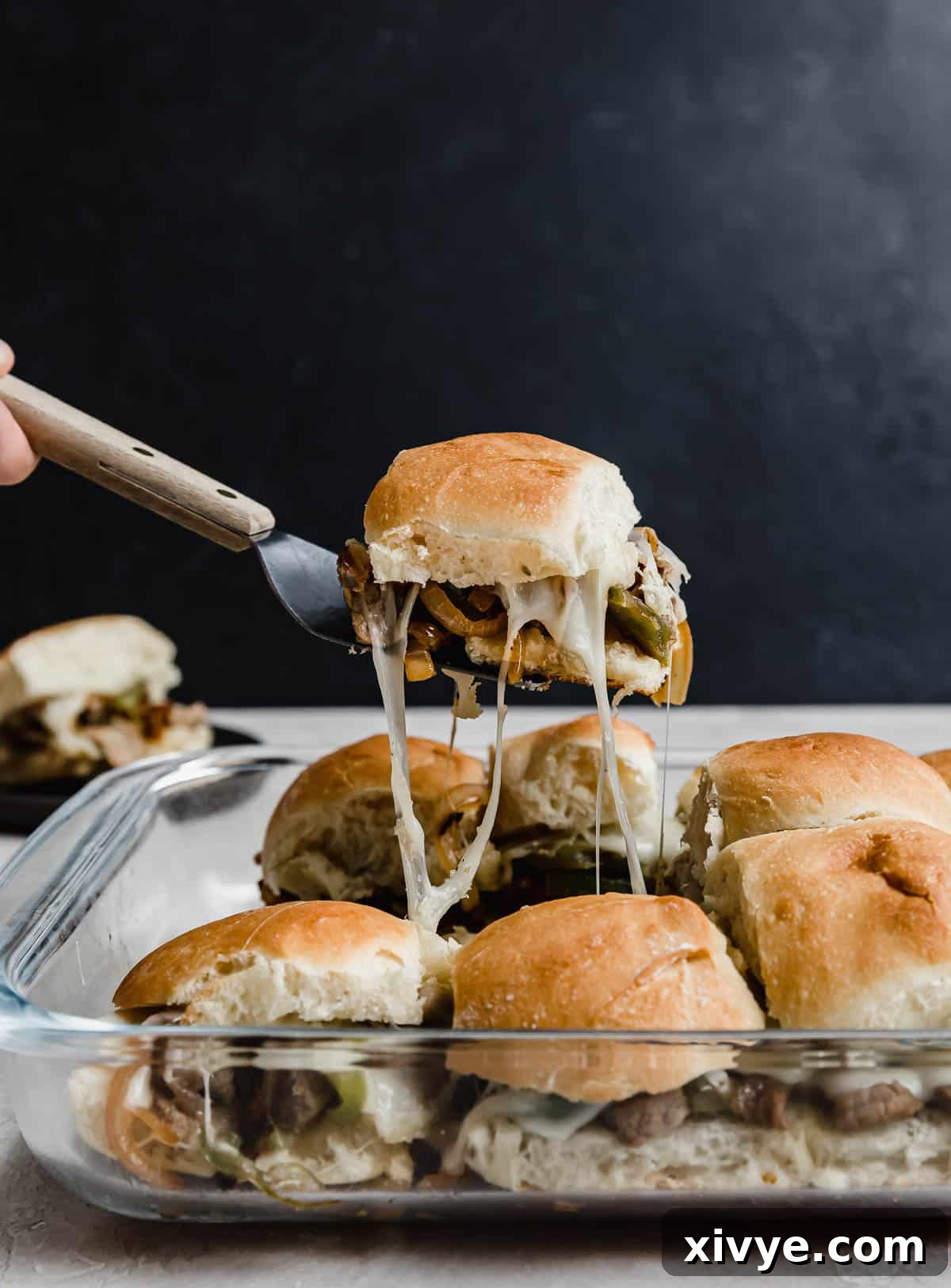 Ultimate Philly Cheesesteak Sliders 8 A Philly Cheesesteak Slider balancing on a metal spatula against a black background with a pan of the best Philly Cheesesteak Sliders recipe in a glass dish beneath the hovering slider.