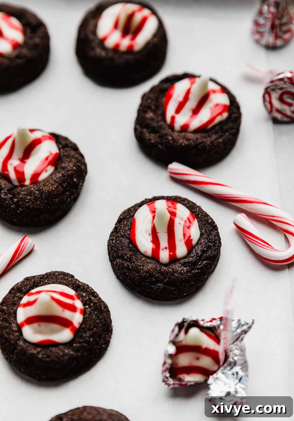 A chocolate cookie topped with a red and white striped peppermint candy kiss, on a white surface with a small candy cane next to the Christmas blossom cookie.