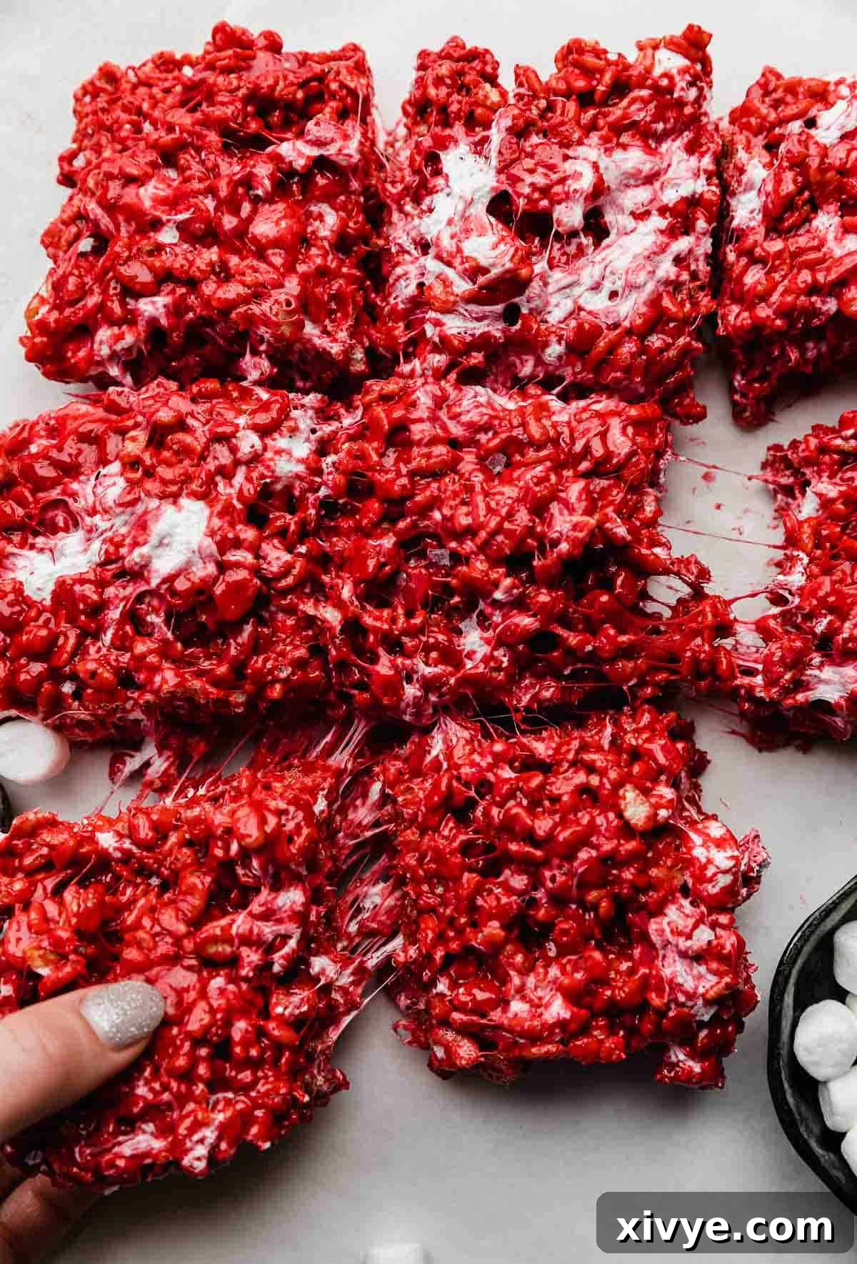 A hand grabbing the a square of bright red gooey Red Velvet Rice Krispies treats.