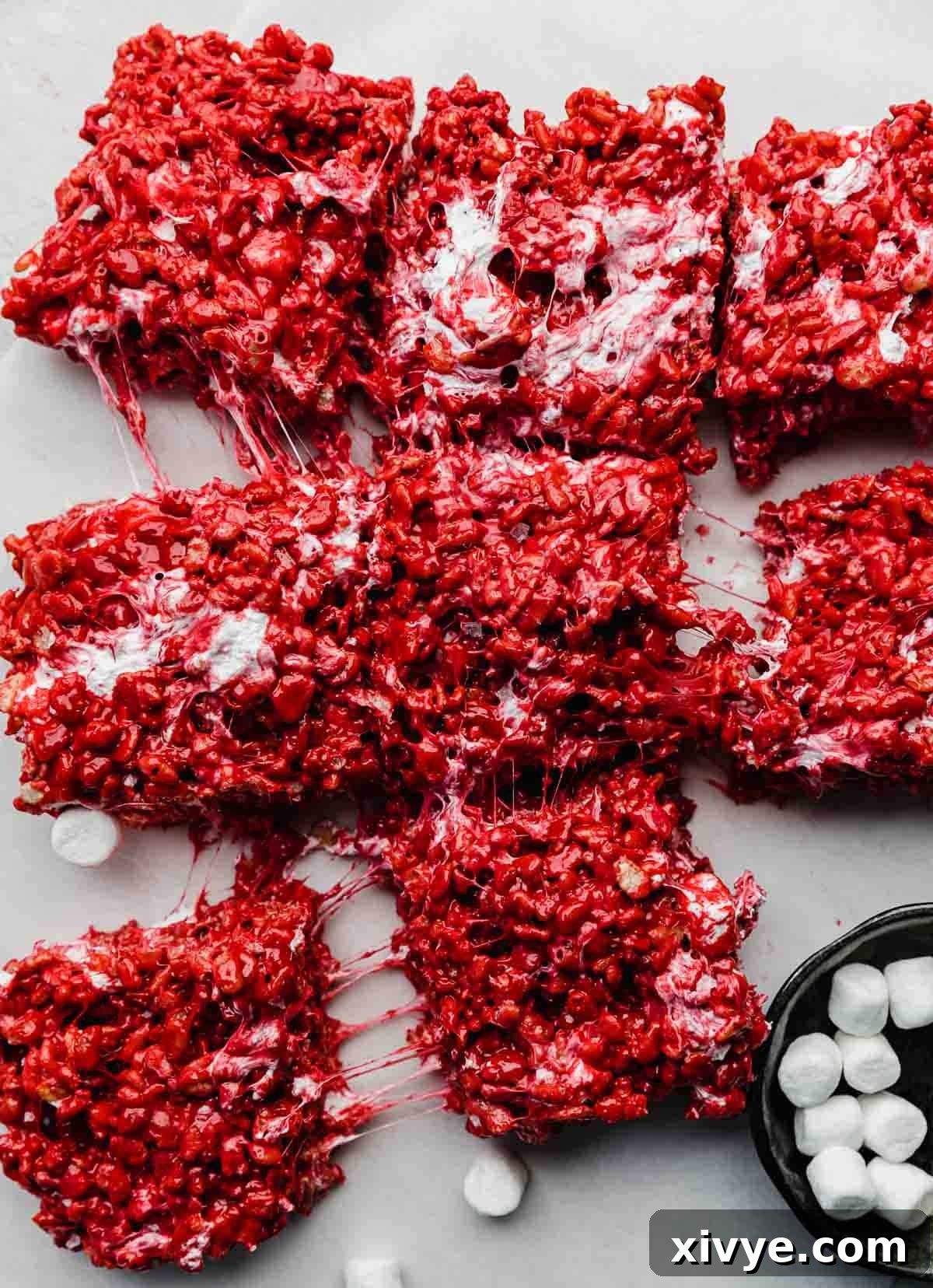 Red Velvet Rice Krispies treats on a light gray background, with the gooey marshmallows showing through as some squares of Rice Krispies are being pulled from the center square.