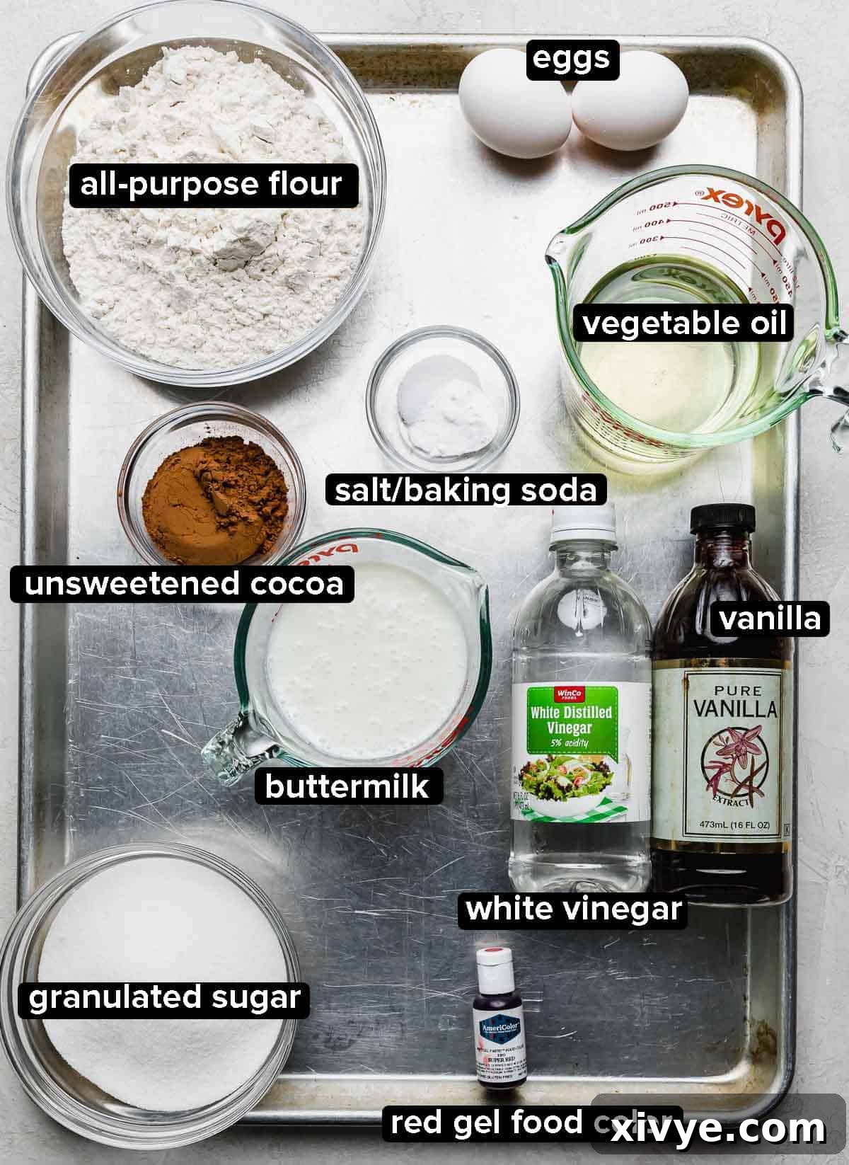 Crimson Crown Bundt 3 Red Velvet Bundt Cake ingredients portioned into glass bowl on a silver baking sheet: oil, buttermilk, vanilla, red gel food color, sugar, cocoa powder, eggs, salt and baking soda, and white vinegar.