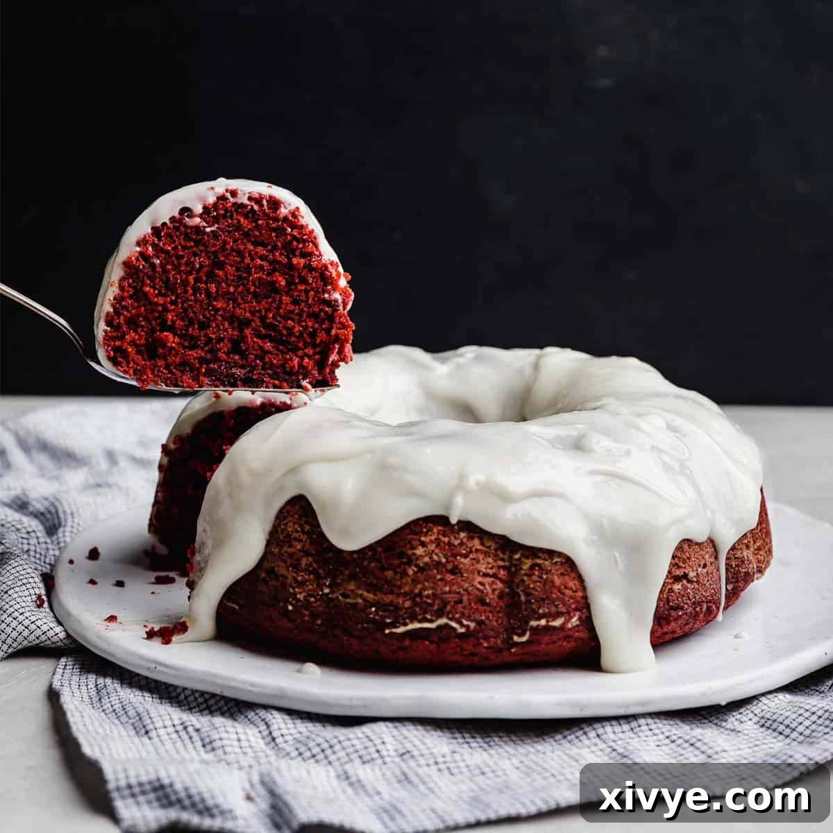Crimson Crown Bundt 7 A slice of red valentines day bundt cake hovering over a cream cheese glaze topped Red Velvet Bundt Cake against a black background.