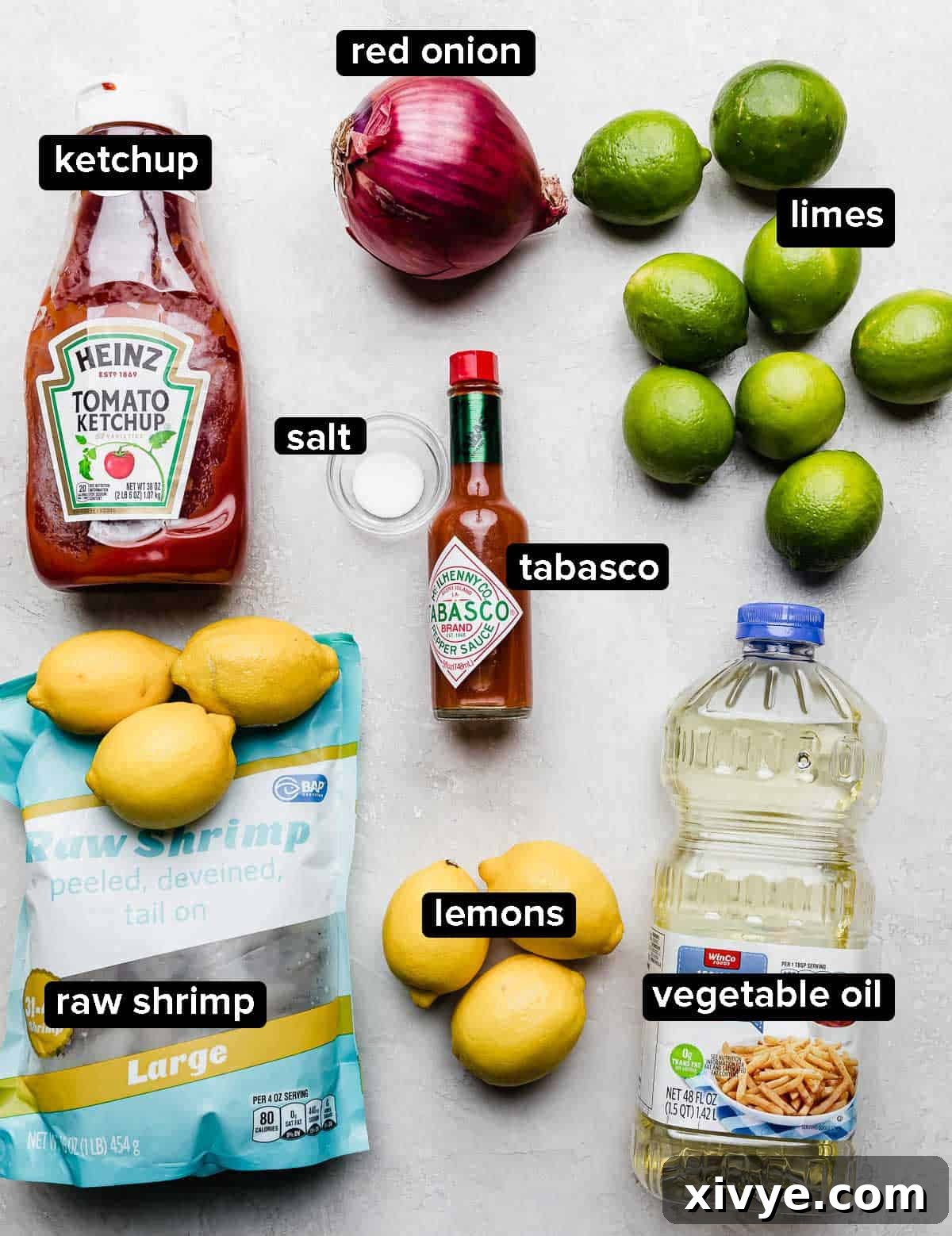 Effortless Cooked Shrimp Ceviche 3 Shrimp Ceviche recipe ingredients laid out on a clean white surface: a bottle of ketchup, a bottle of oil, fresh lemons and limes, Tabasco sauce, raw shrimp, and a small bowl of salt.