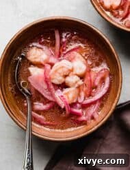 Effortless Cooked Shrimp Ceviche 8 Shrimp Ceviche with sliced purple onions in a ketchup and tabasco based lemon lime sauce, in a bowl on a light gray surface.