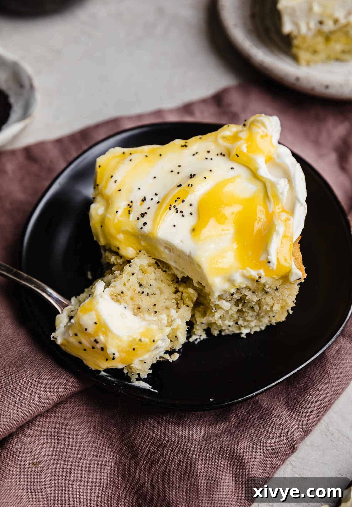 A square slice of Lemon and Poppy Seed Cake topped with whipped cream cheese frosting and lemon curd on a black plate.