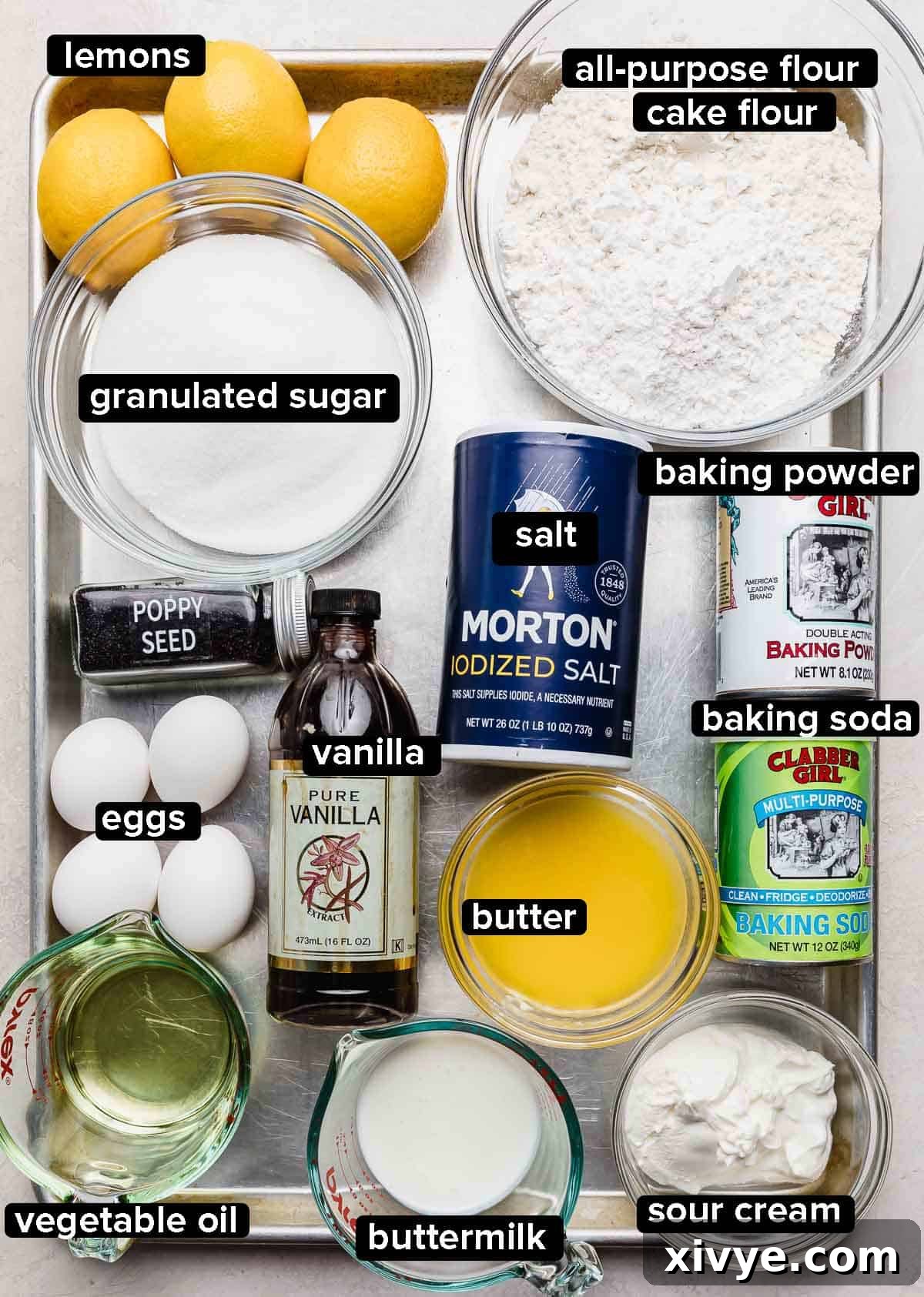 Lemon and Poppy Seed Cake ingredients on a silver baking sheet: sugar, flour, salt, eggs, oil, butter, leaning agents, sour cream, and real lemons.
