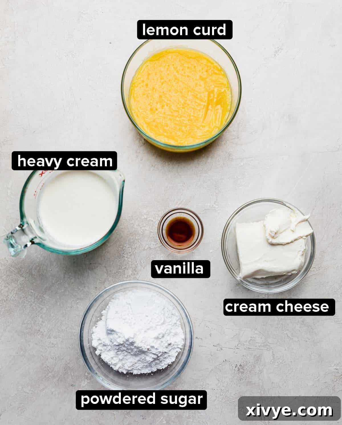 Ingredients used to make a whipped cream cheese frosting with lemon curd on a light gray surface: heavy cream, powdered sugar, cream cheese, vanilla, and lemon curd.