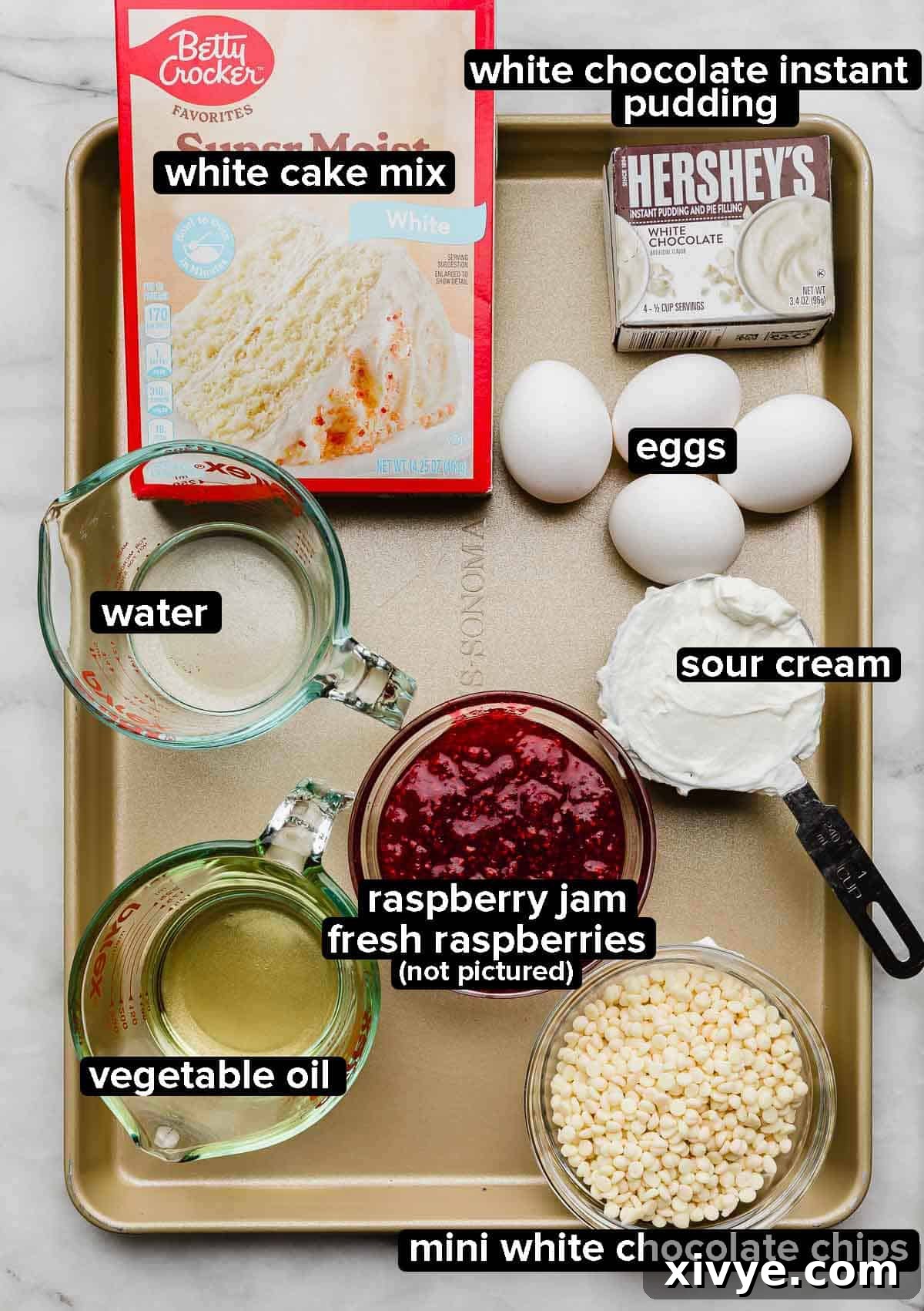 White Chocolate Raspberry Bundt Cake recipe ingredients portioned into glass bowls on a bronze baking sheet: white cake mix, white chocolate pudding, white chocolate chips, raspberry jam, oil, water, eggs, and sour cream.
