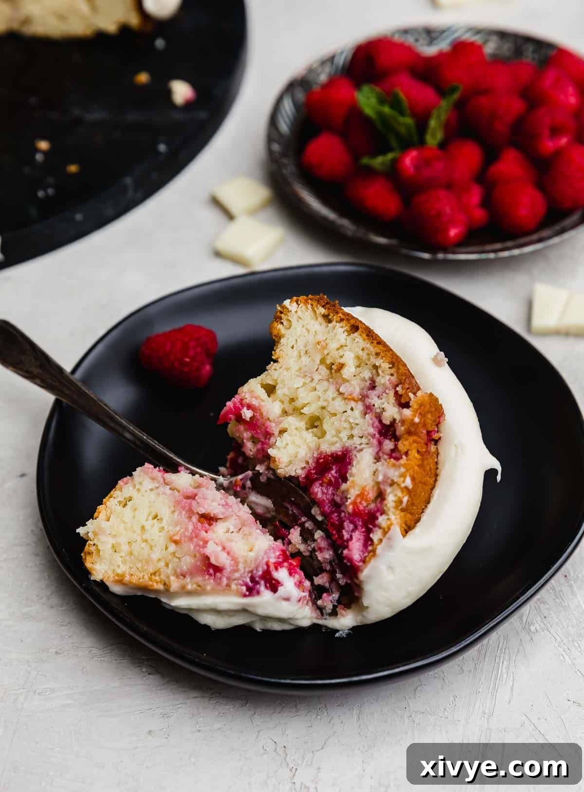 A slice of White Chocolate Raspberry Bundt Cake topped with cream cheese frosting, with a slice on a black plate.